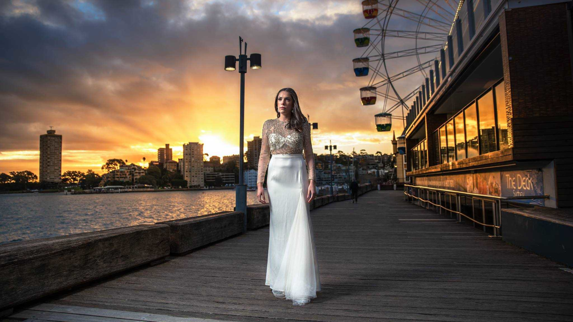 Bride in an elegant gown stands on a harbor boardwalk at sunset with city skyline in the background.