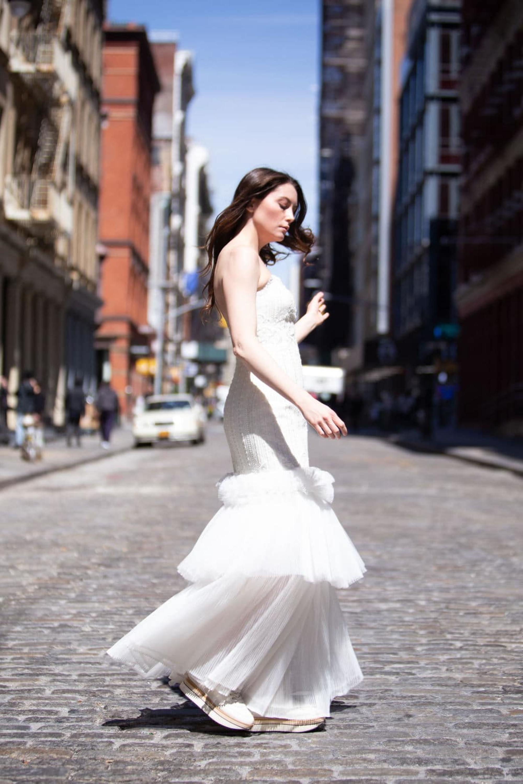 Bride in a fitted mermaid wedding dress walking along a sunlit cobblestone city street.