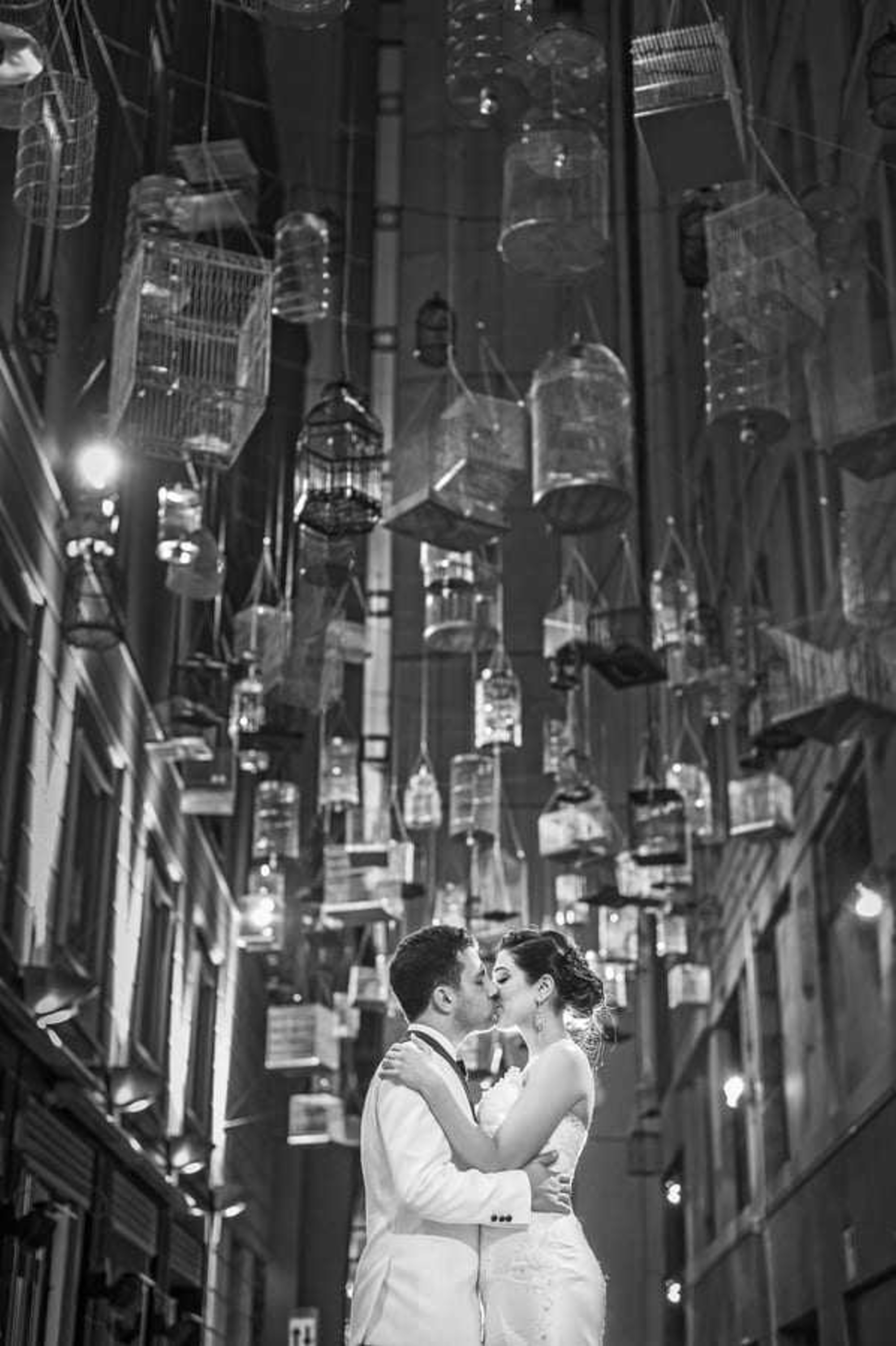Bride and groom embrace and kiss in a narrow city alley under hanging birdcages at night.