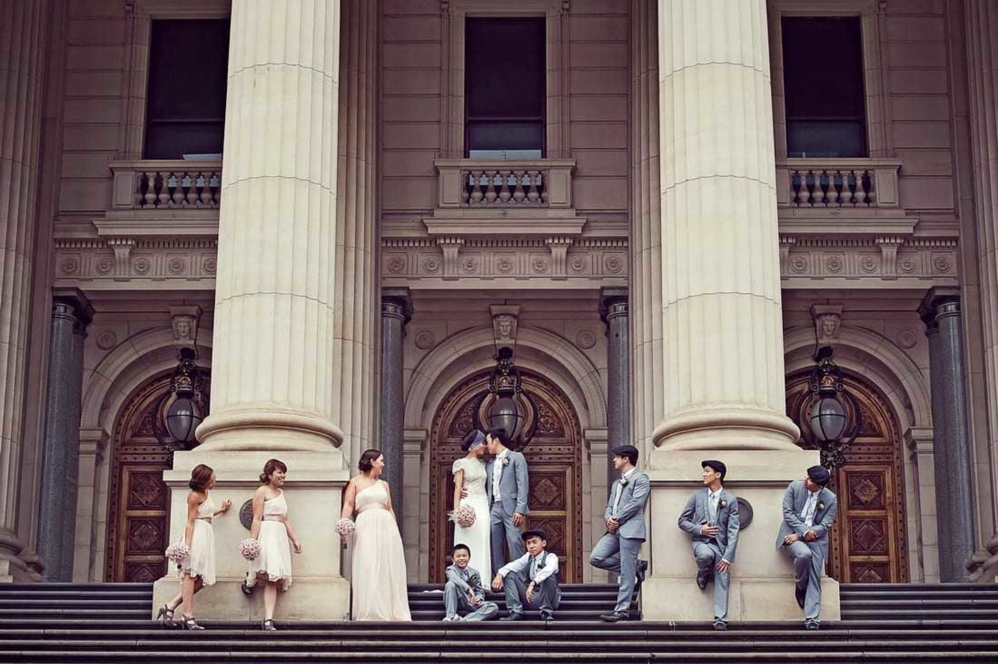 Wedding party poses on grand stone steps in front of a historic building with tall columns.