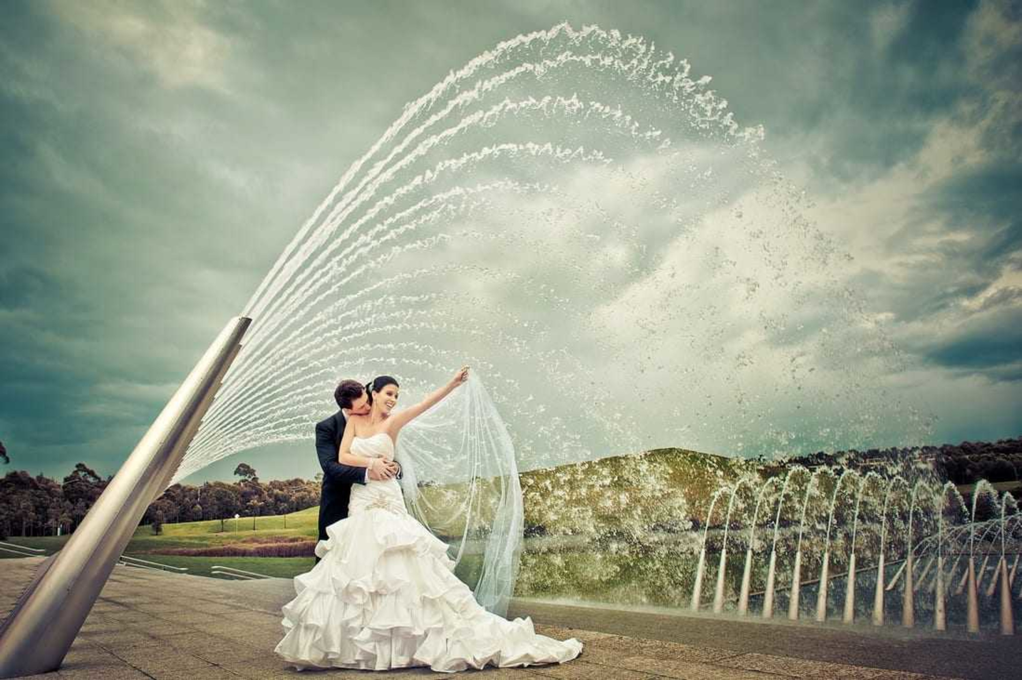 Bride and groom embrace in front of dramatic arcing fountains in a scenic outdoor setting.