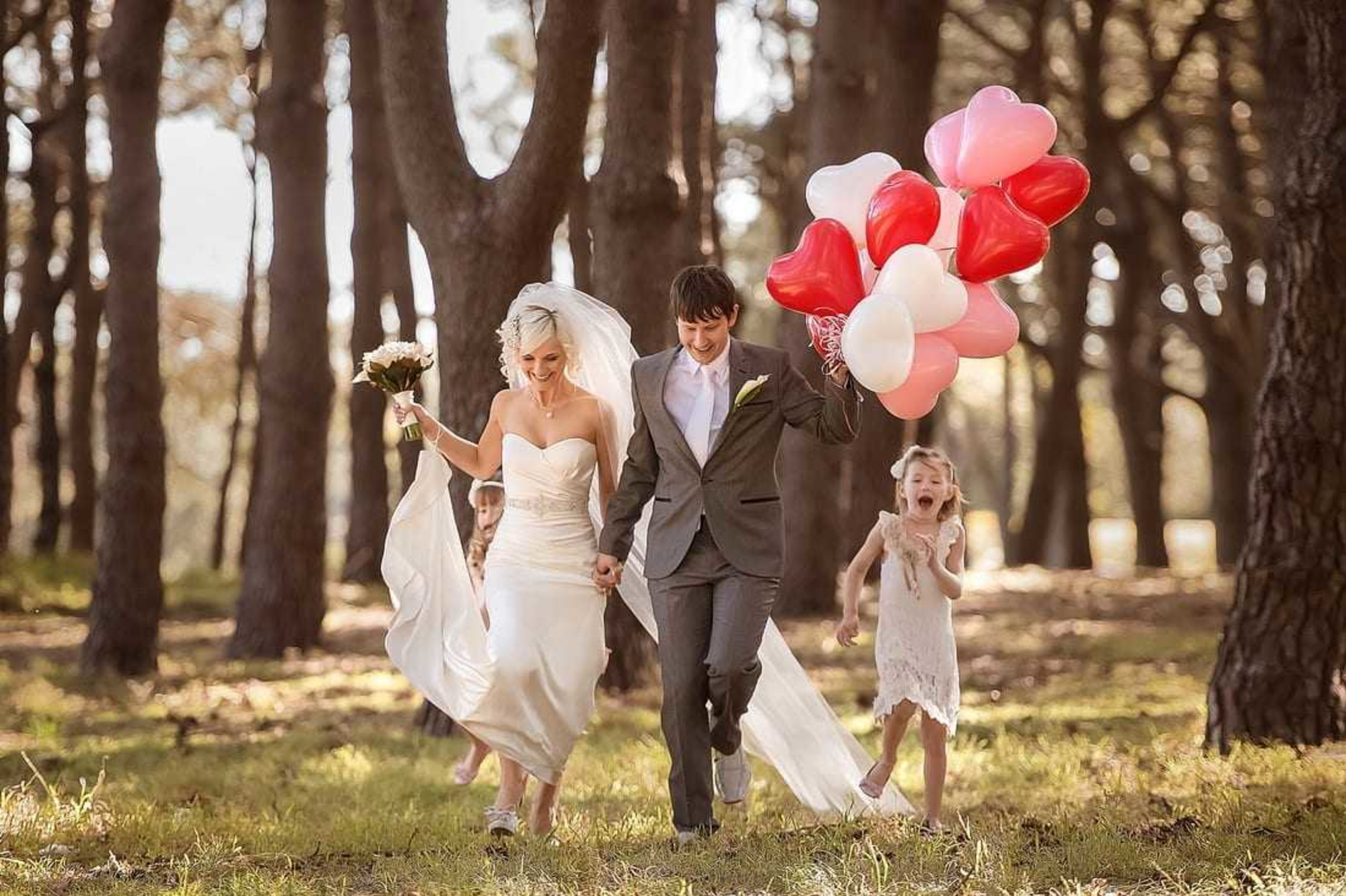 Bride and groom run through a sunlit forest holding heart balloons with joyful children behind them.
