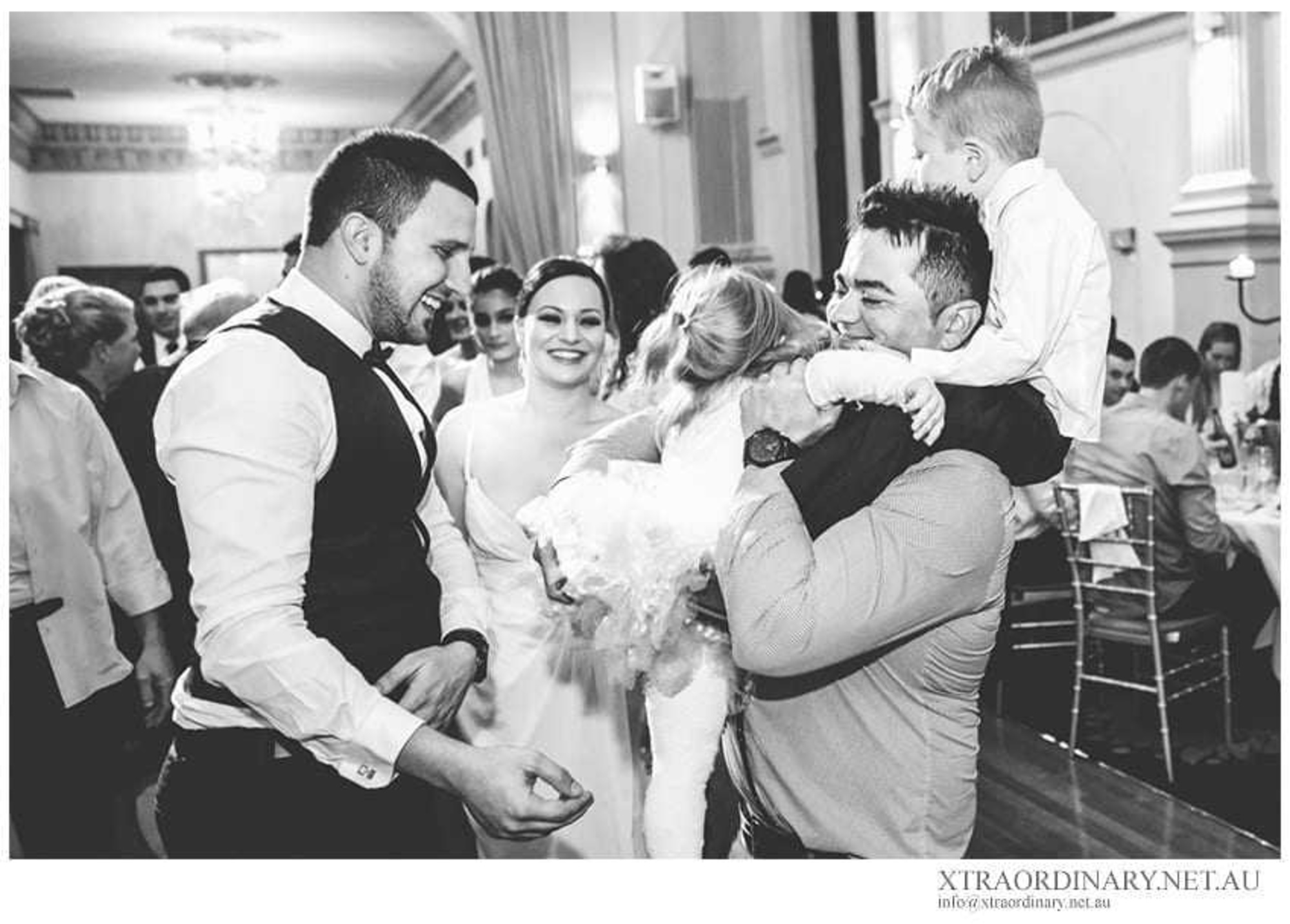 Candid black and white photo of a bride and guests laughing and dancing with children at a lively wedding reception.