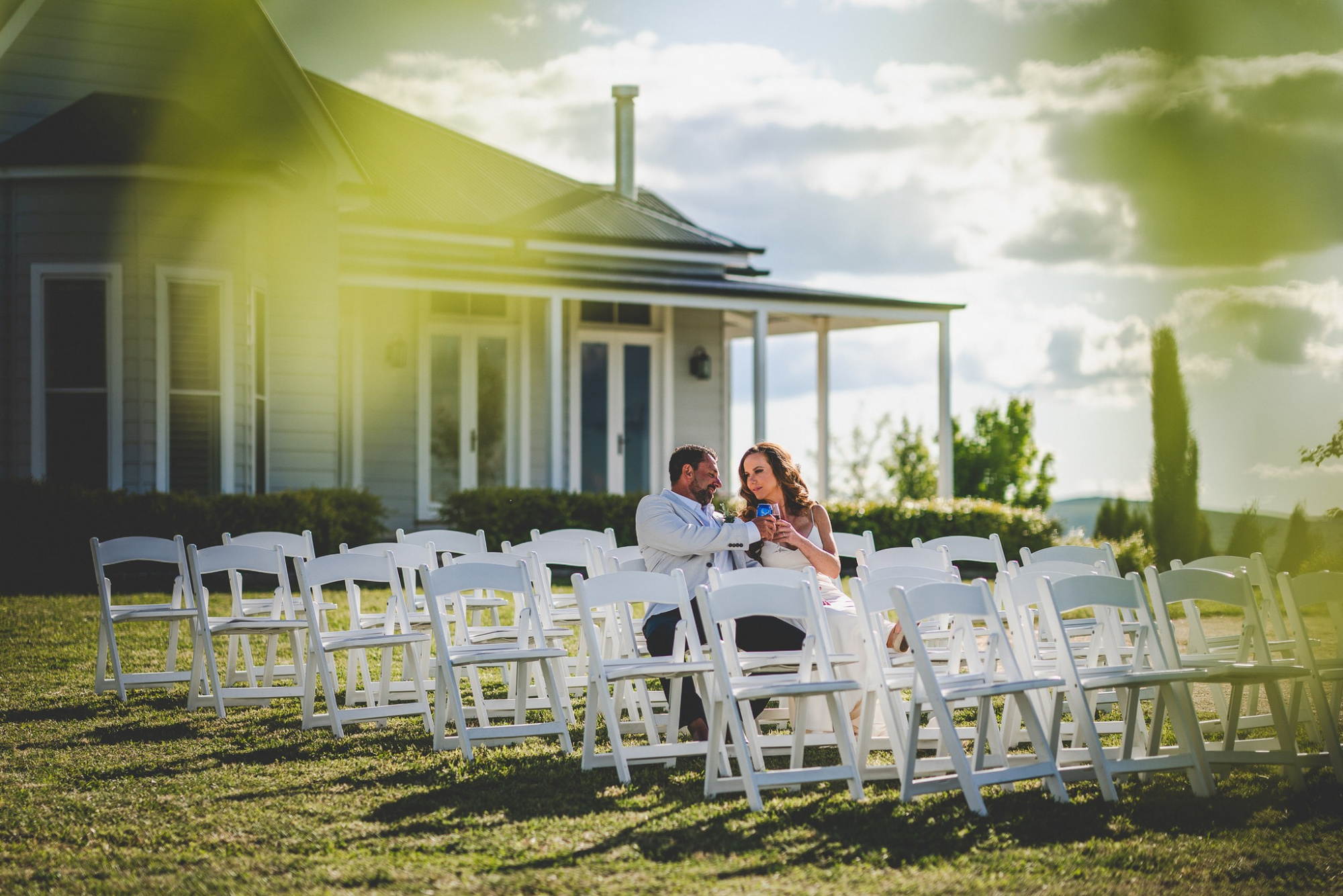 A couple sits together among white ceremony chairs on the lawn of a country house wedding venue at sunset.