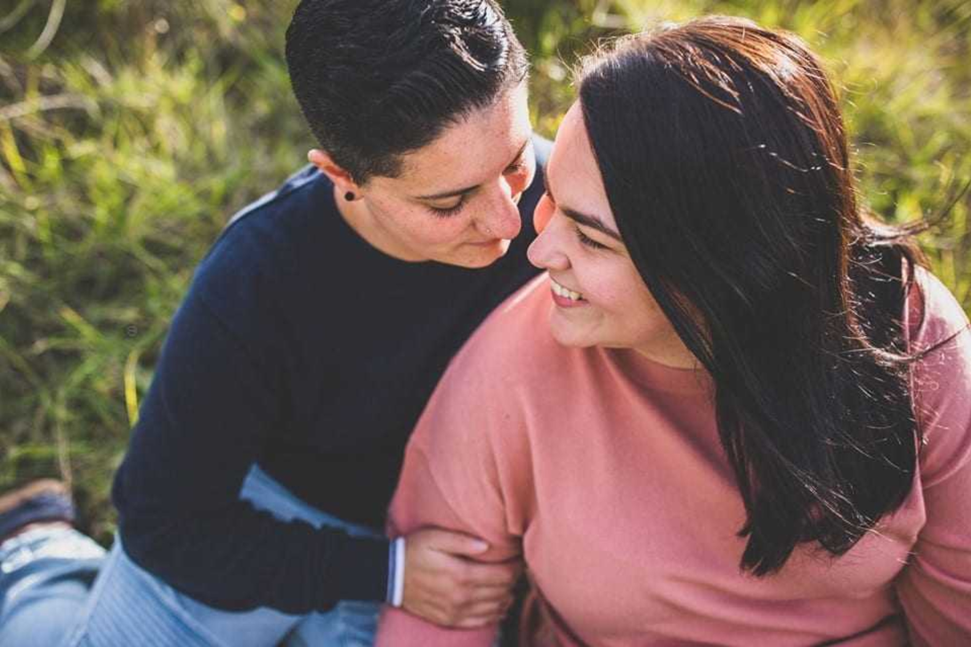 A couple sits closely together in a grassy field, smiling and sharing an intimate moment.