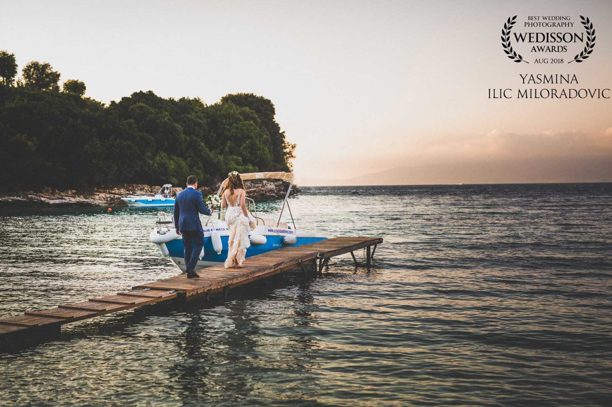 Bride and groom walk along a wooden dock toward a small boat on the water at sunset.
