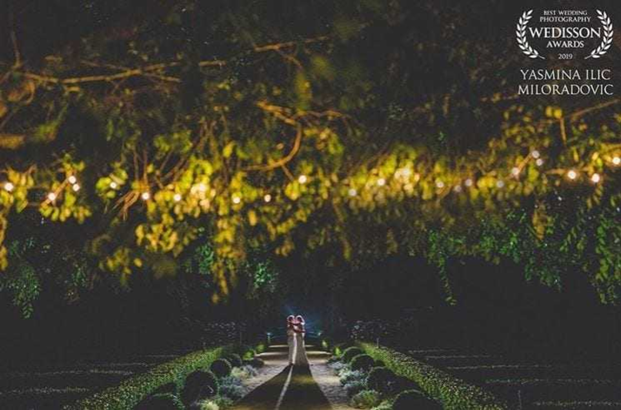 Newlywed couple embraces on a garden path at night beneath glowing string lights and overhanging trees.