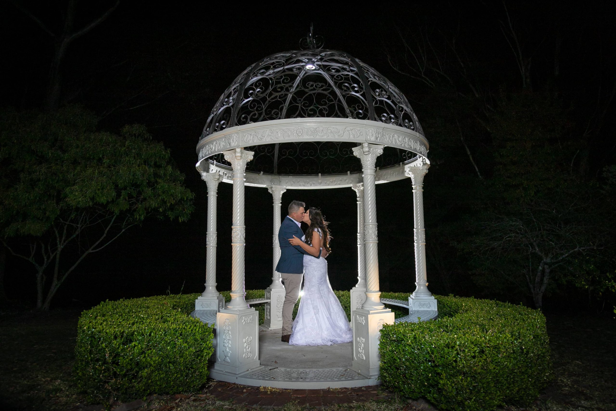 A couple kisses under a lit white gazebo in a garden at night.