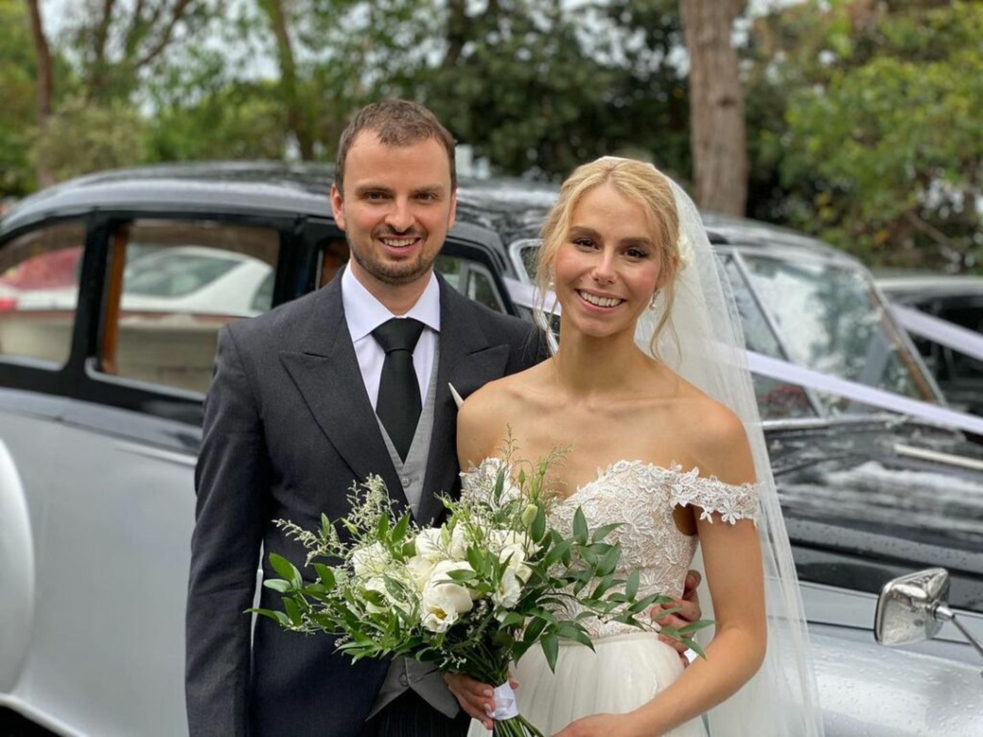 Bride and groom smiling in front of a classic car, holding a white and green bridal bouquet.