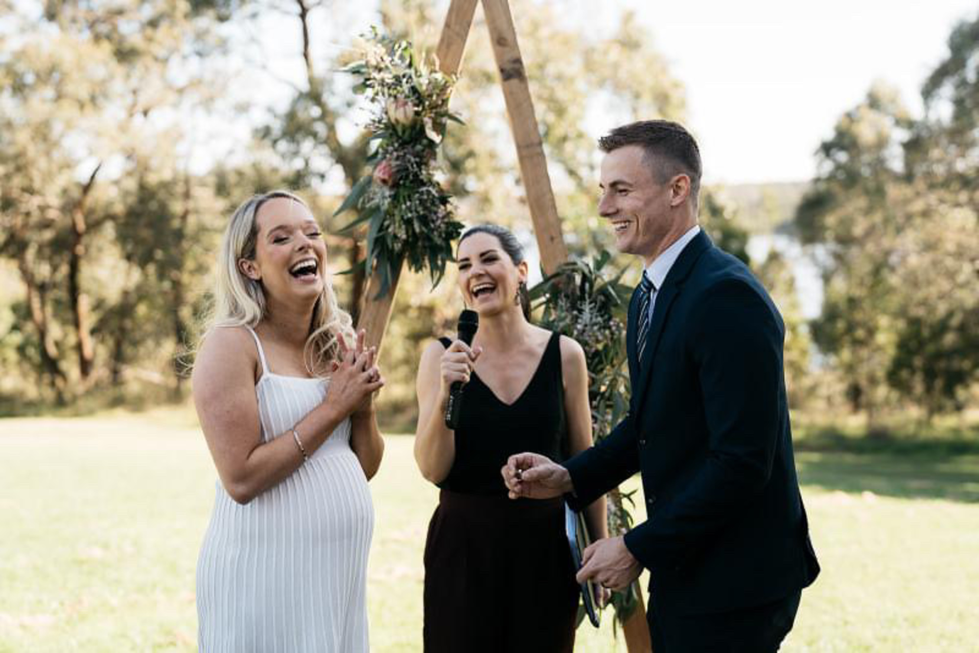 A joyful couple laughs with their celebrant during an outdoor wedding ceremony under a rustic floral arch.