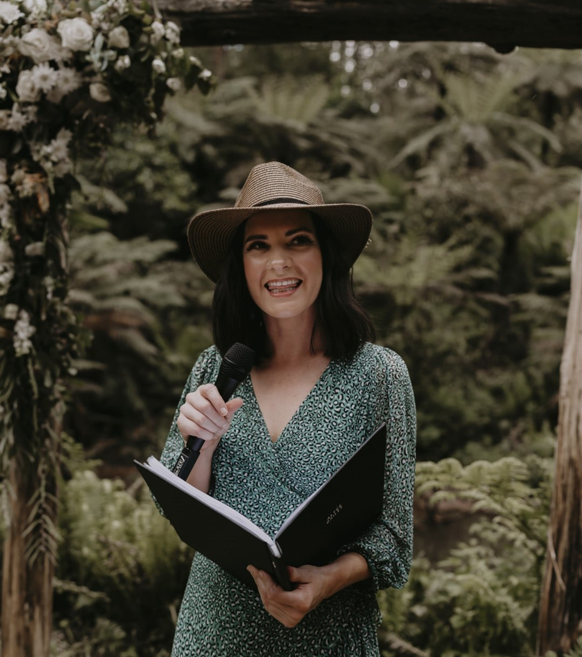 Wedding celebrant in a hat holding a microphone and ceremony book in a lush outdoor garden setting.