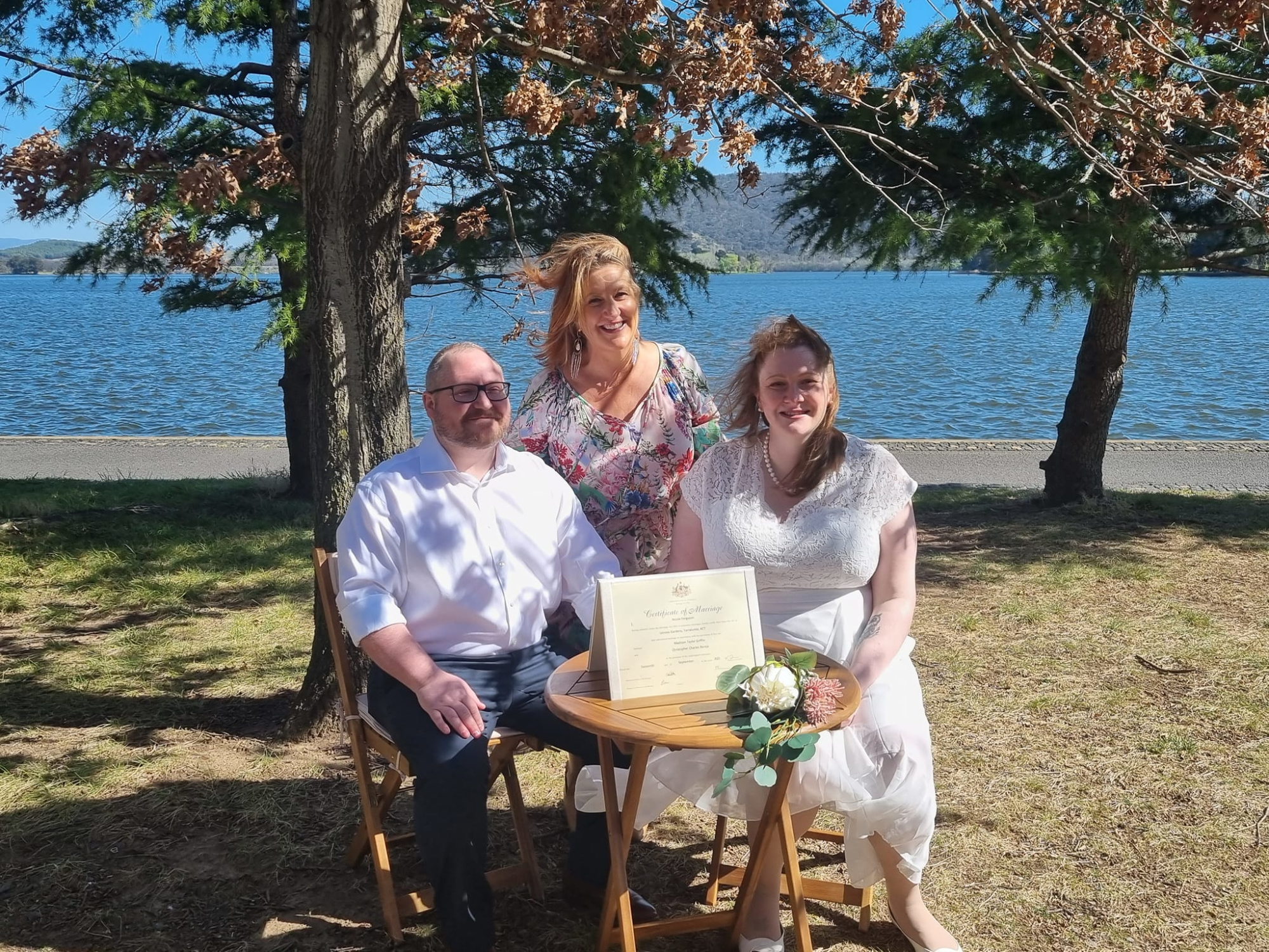 Newly married couple with their celebrant posing by a lakeside table holding the marriage certificate.