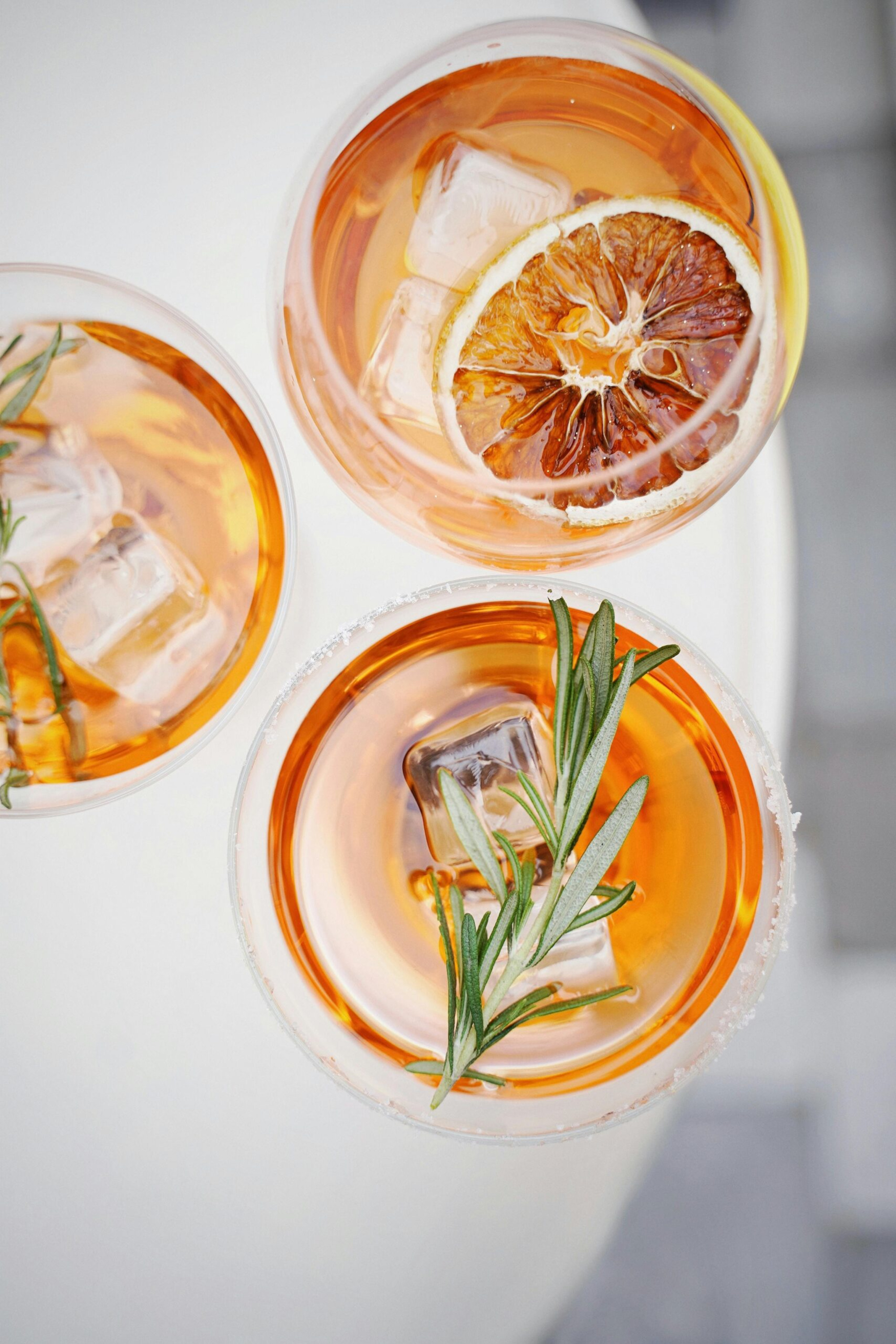 Overhead view of three amber cocktails with ice, citrus, and rosemary garnishes on a light surface.