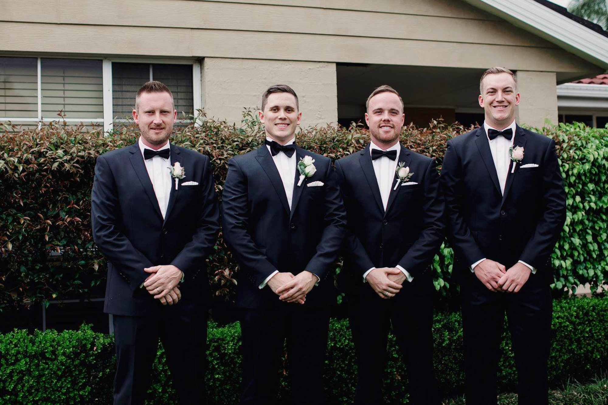 Four groomsmen in black tuxedos with boutonnieres pose together in a garden outside a house.