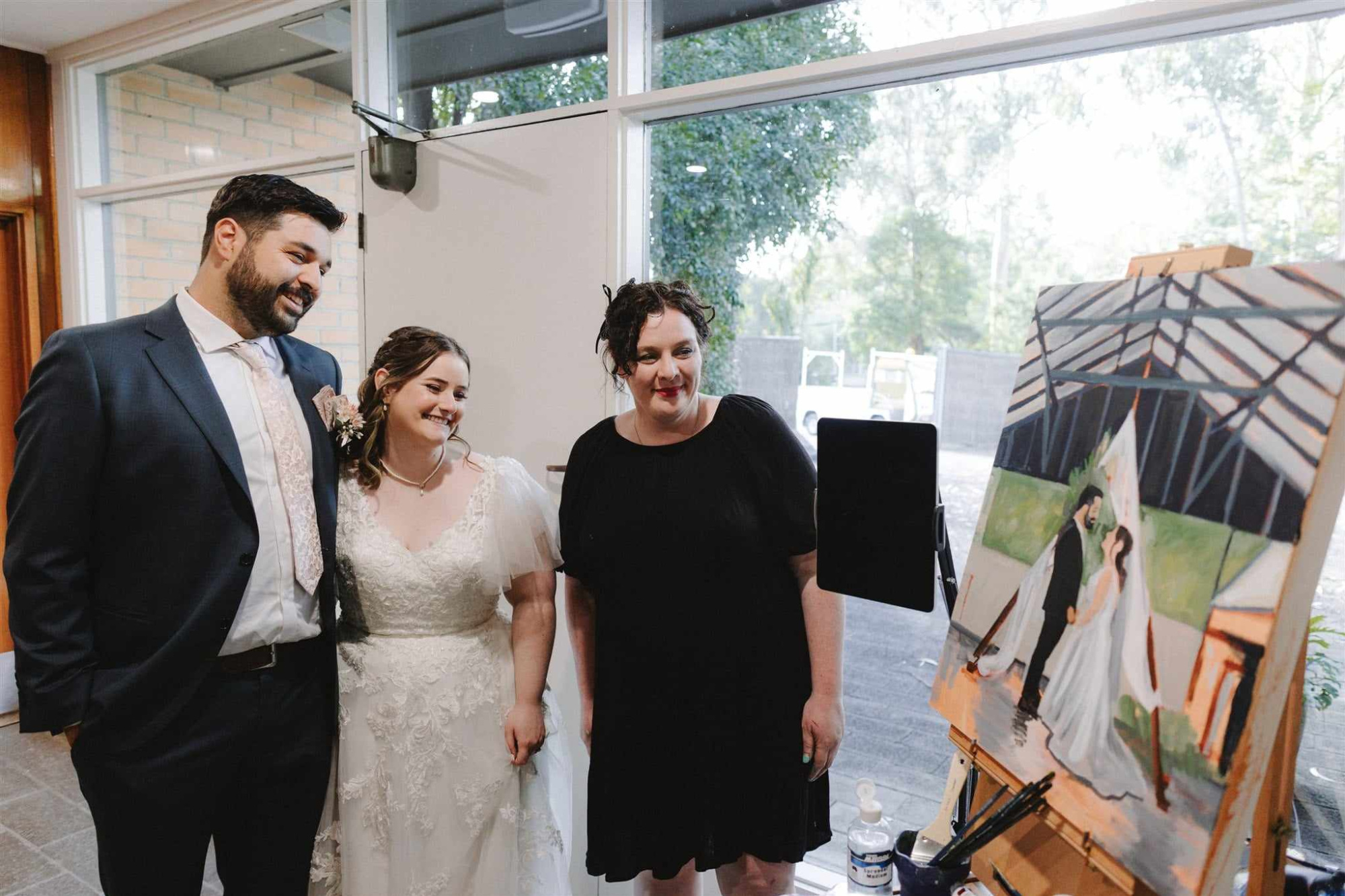 Bride and groom smile with a live wedding painter beside an easel displaying their ceremony painting.