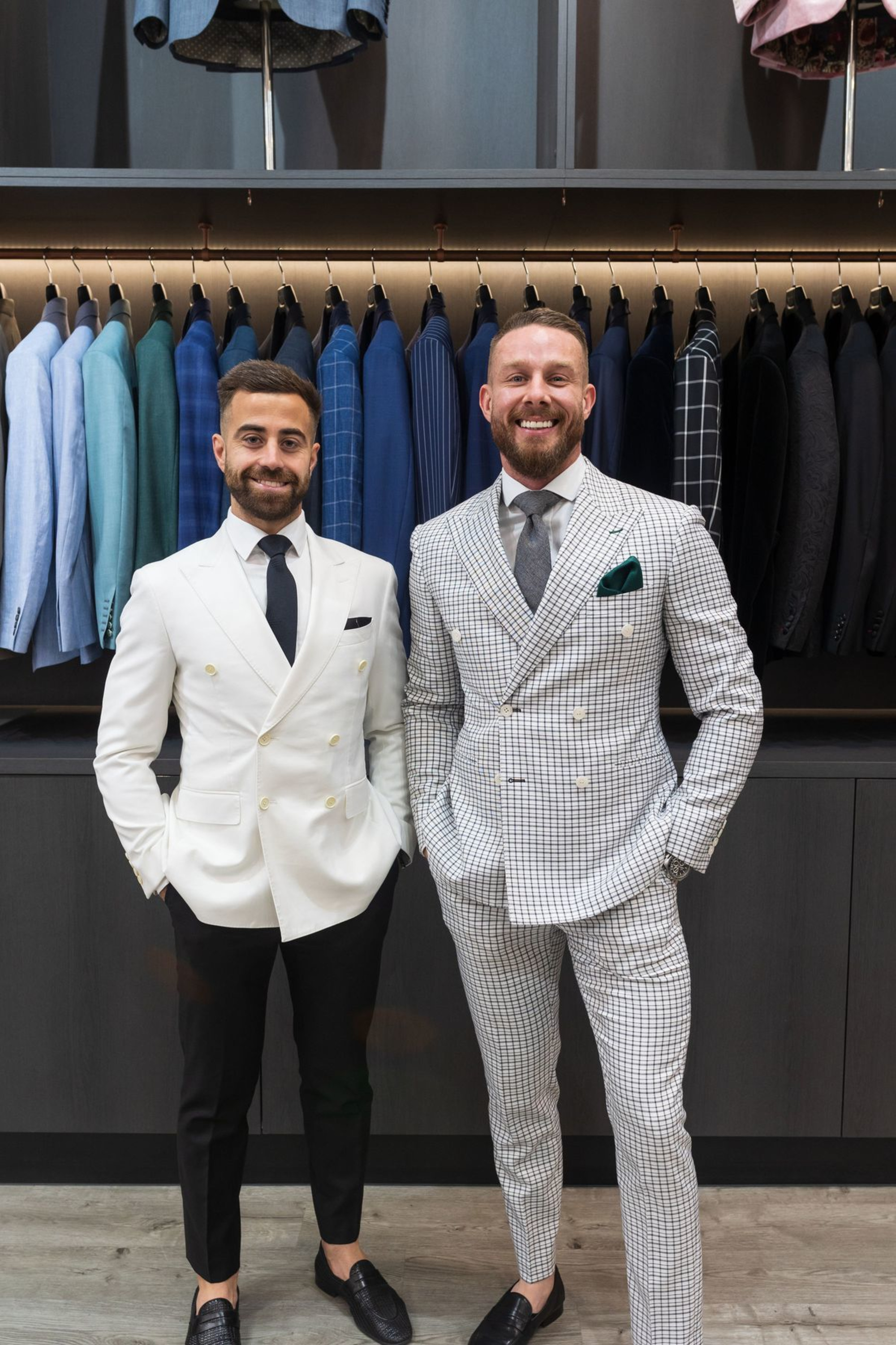 Two men in stylish suits stand in a menswear shop with rows of colorful jackets behind them.