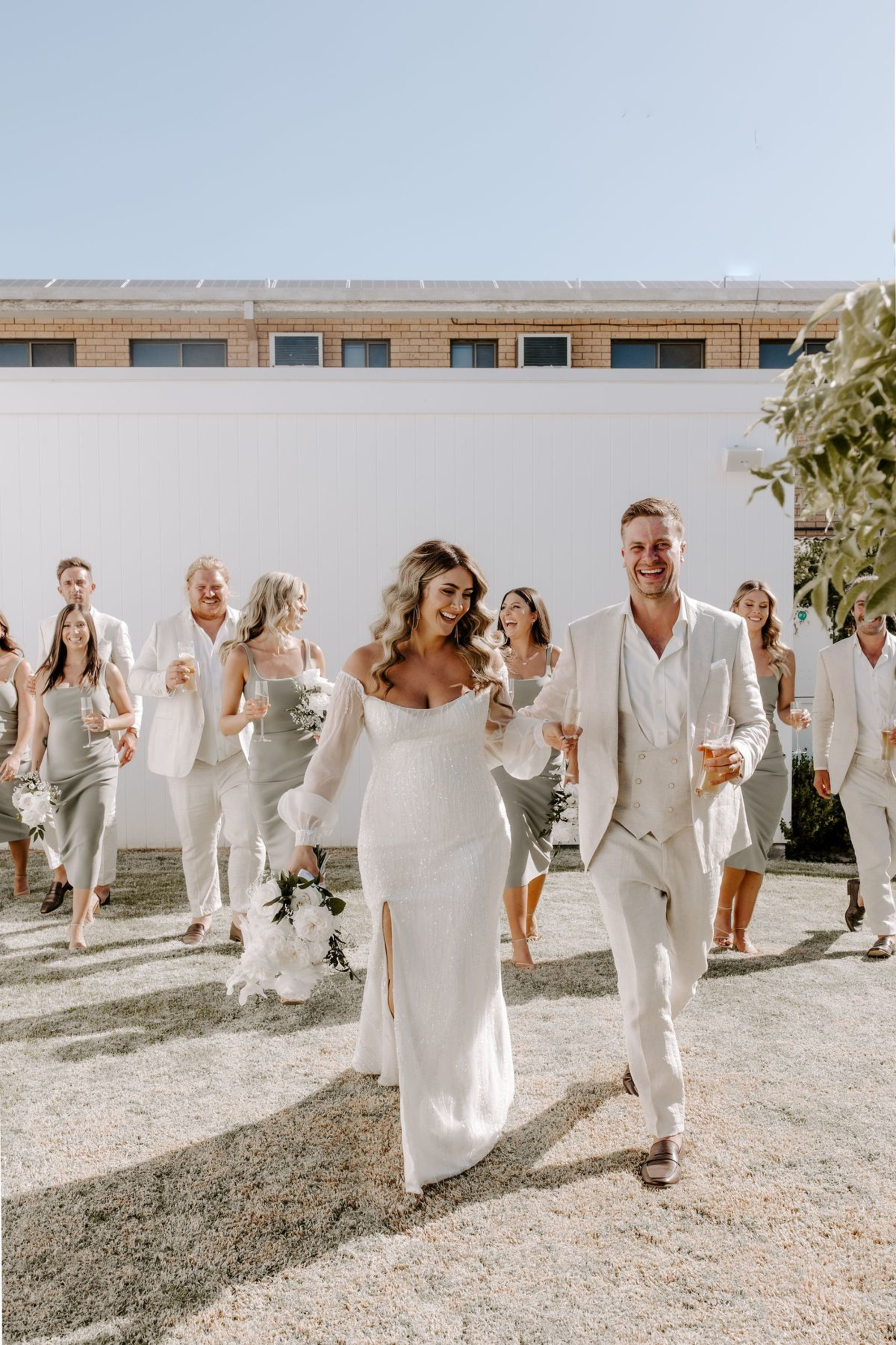 Bride and groom walk with their bridal party outdoors in neutral-toned attire, holding champagne and white bouquets.