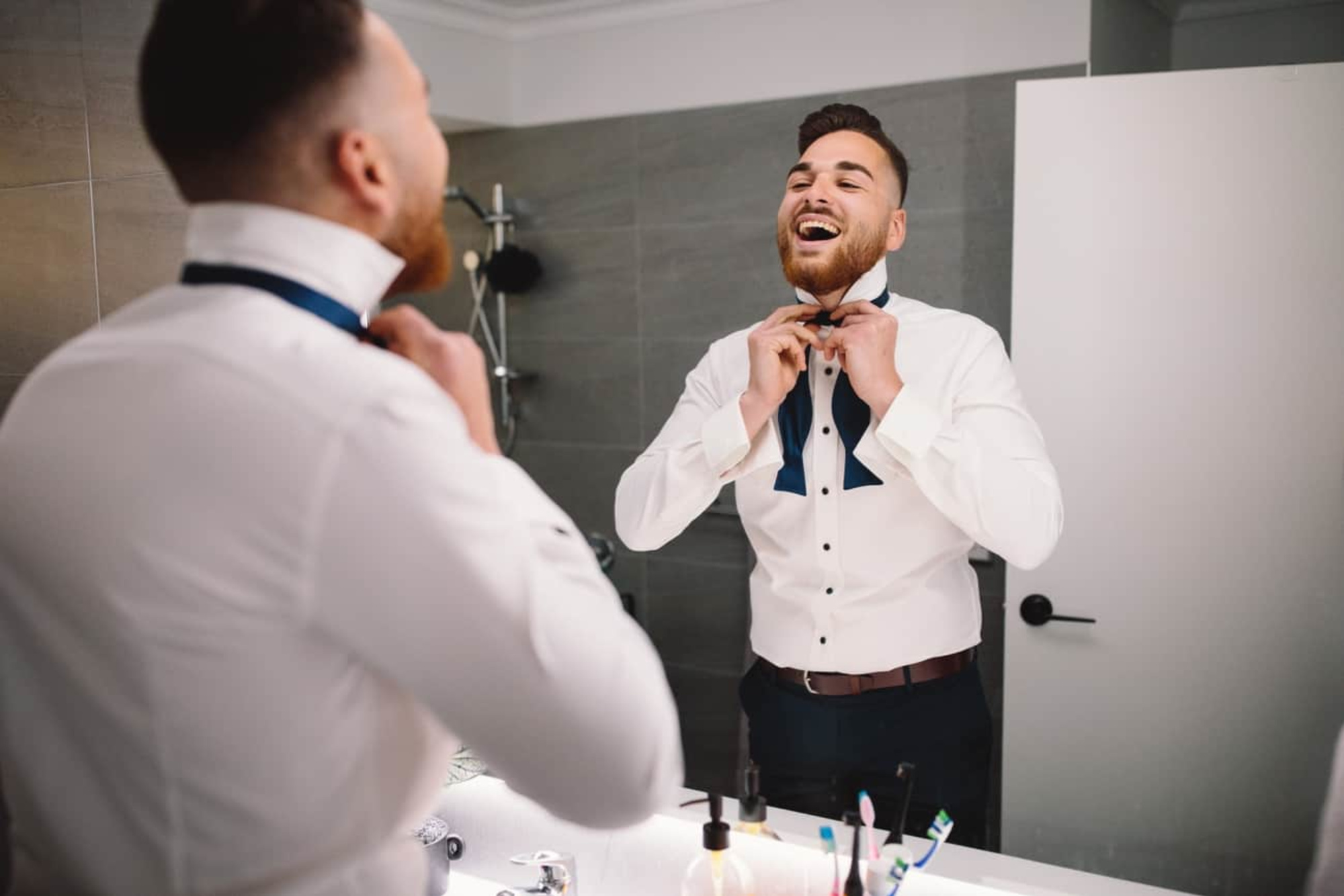 Groom smiling in the bathroom mirror while adjusting his navy bow tie before the wedding.