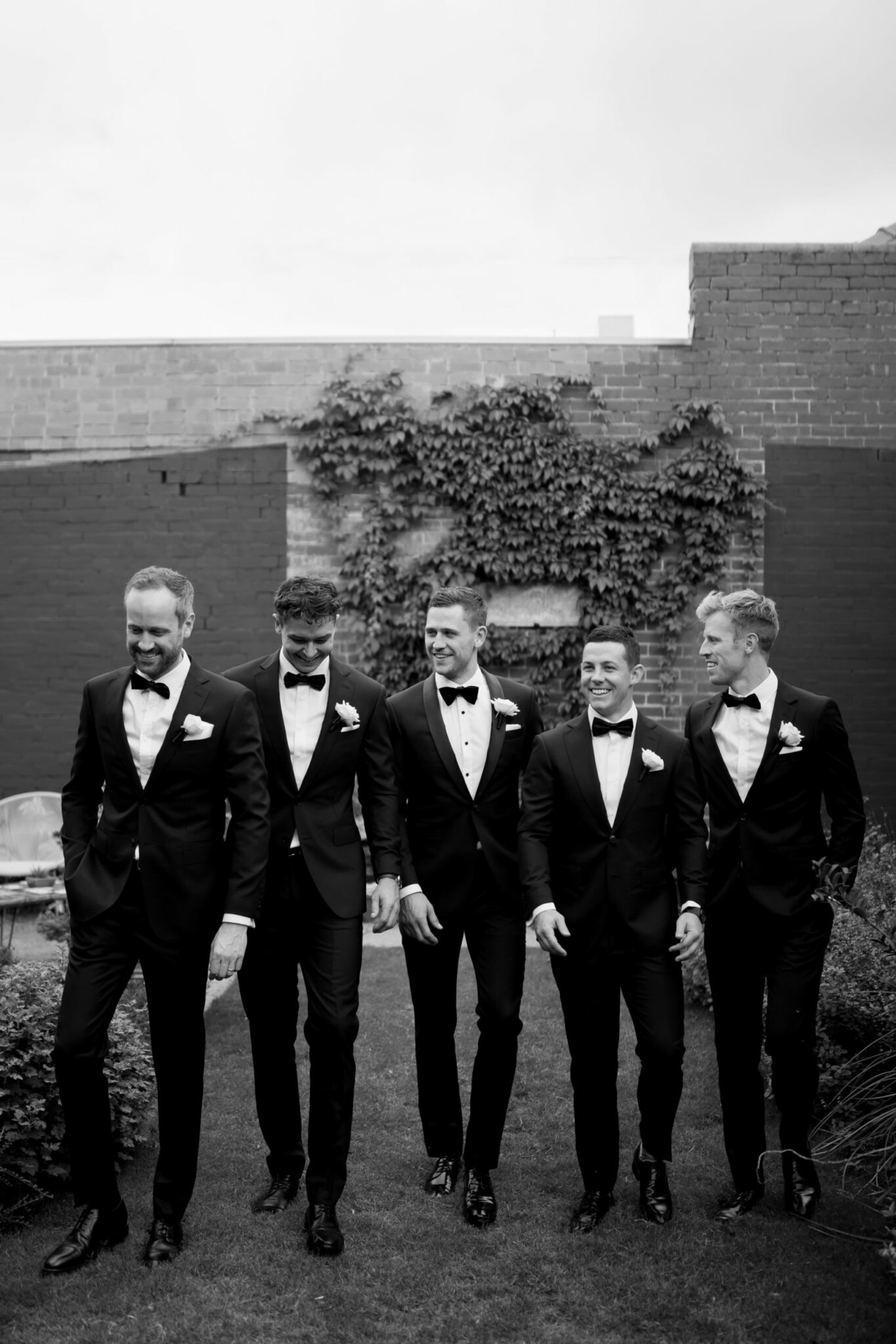Five groomsmen in black tuxedos walk together in a garden courtyard with a brick wall and ivy backdrop.