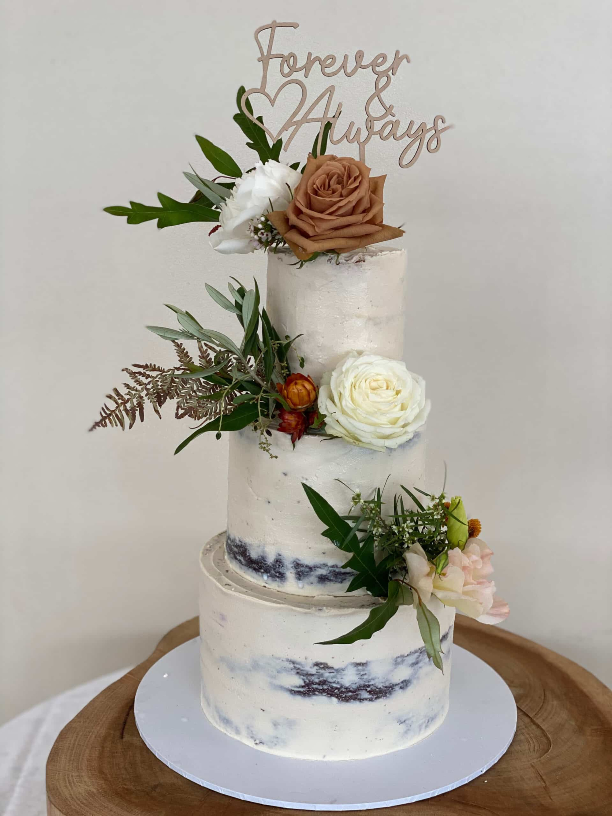Three-tier semi-naked wedding cake with floral decorations and a Forever & Always topper on a wooden stand.