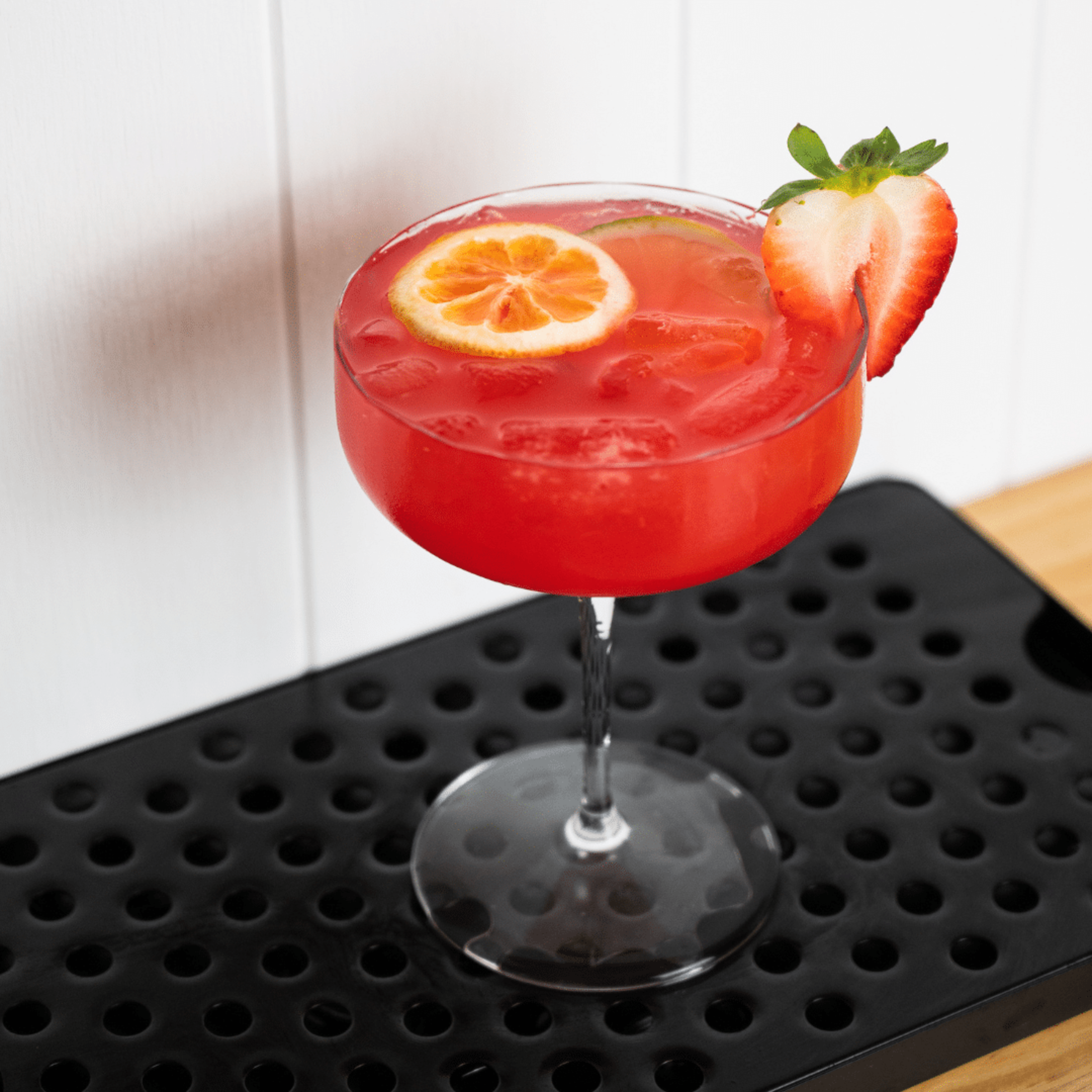 Bright red strawberry citrus cocktail in a coupe glass on a black bar mat.