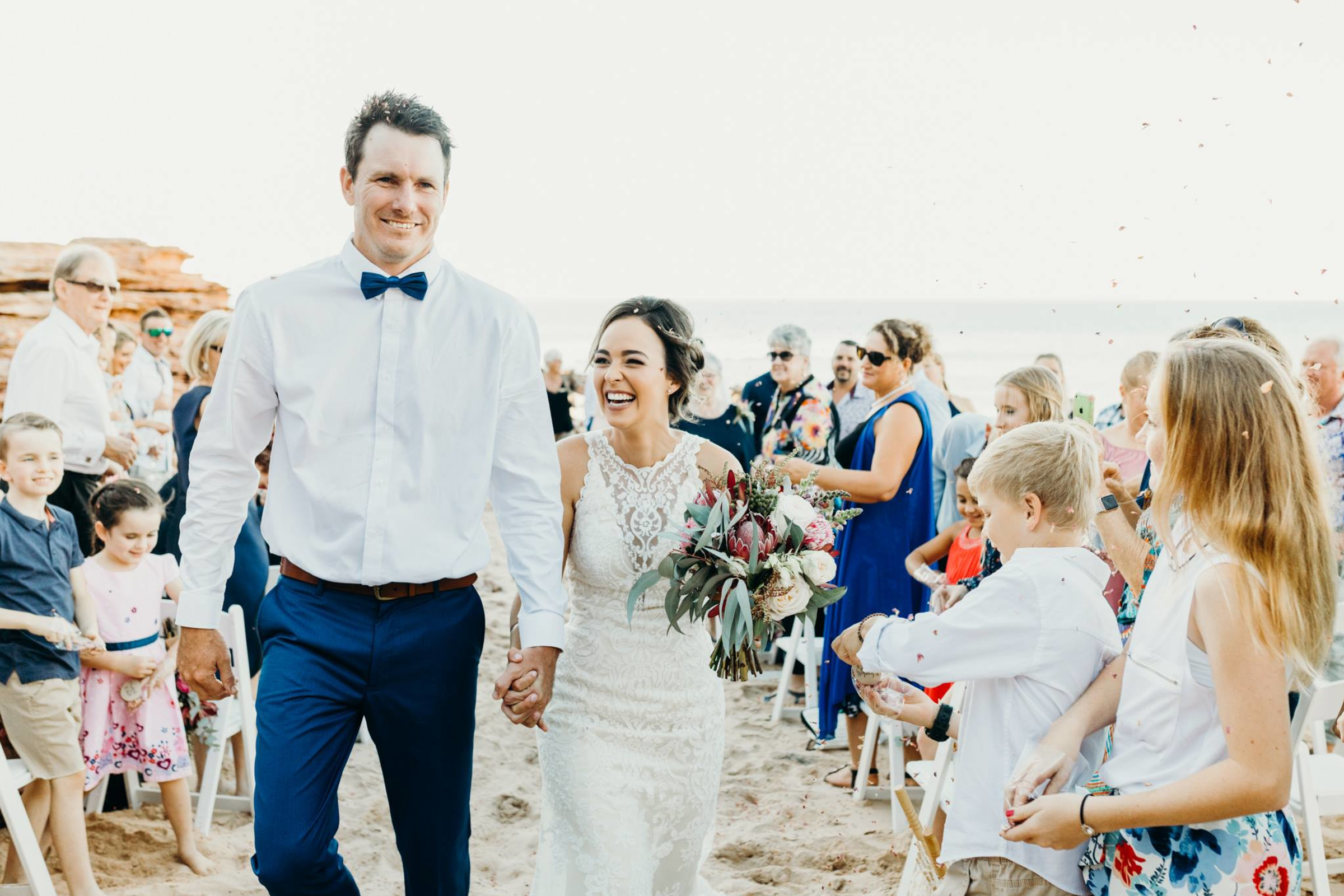 A joyful couple walks hand in hand down a sandy beach aisle as guests celebrate around them.