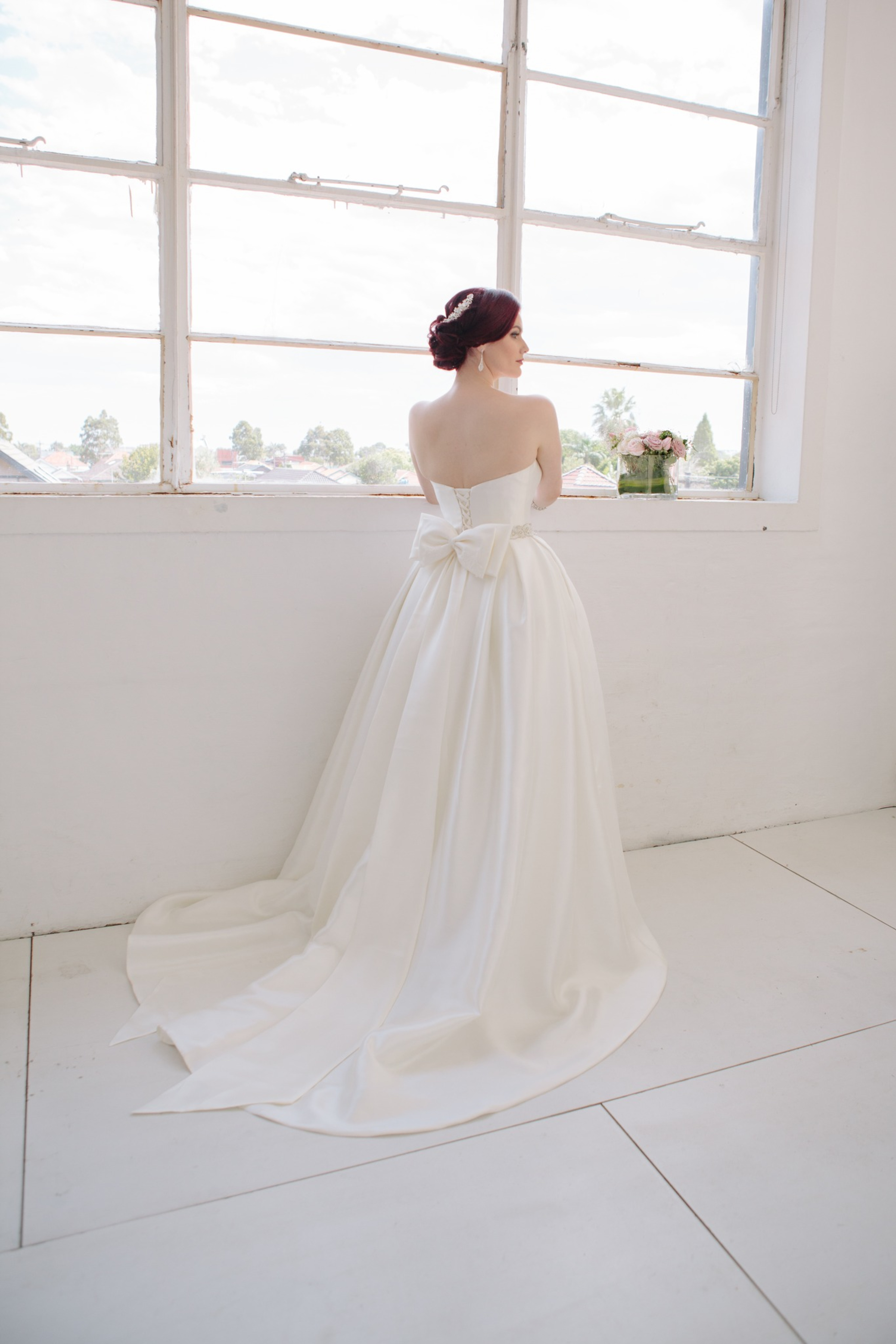 Bride in a strapless ballgown with a large bow train stands by a bright window holding a bouquet.