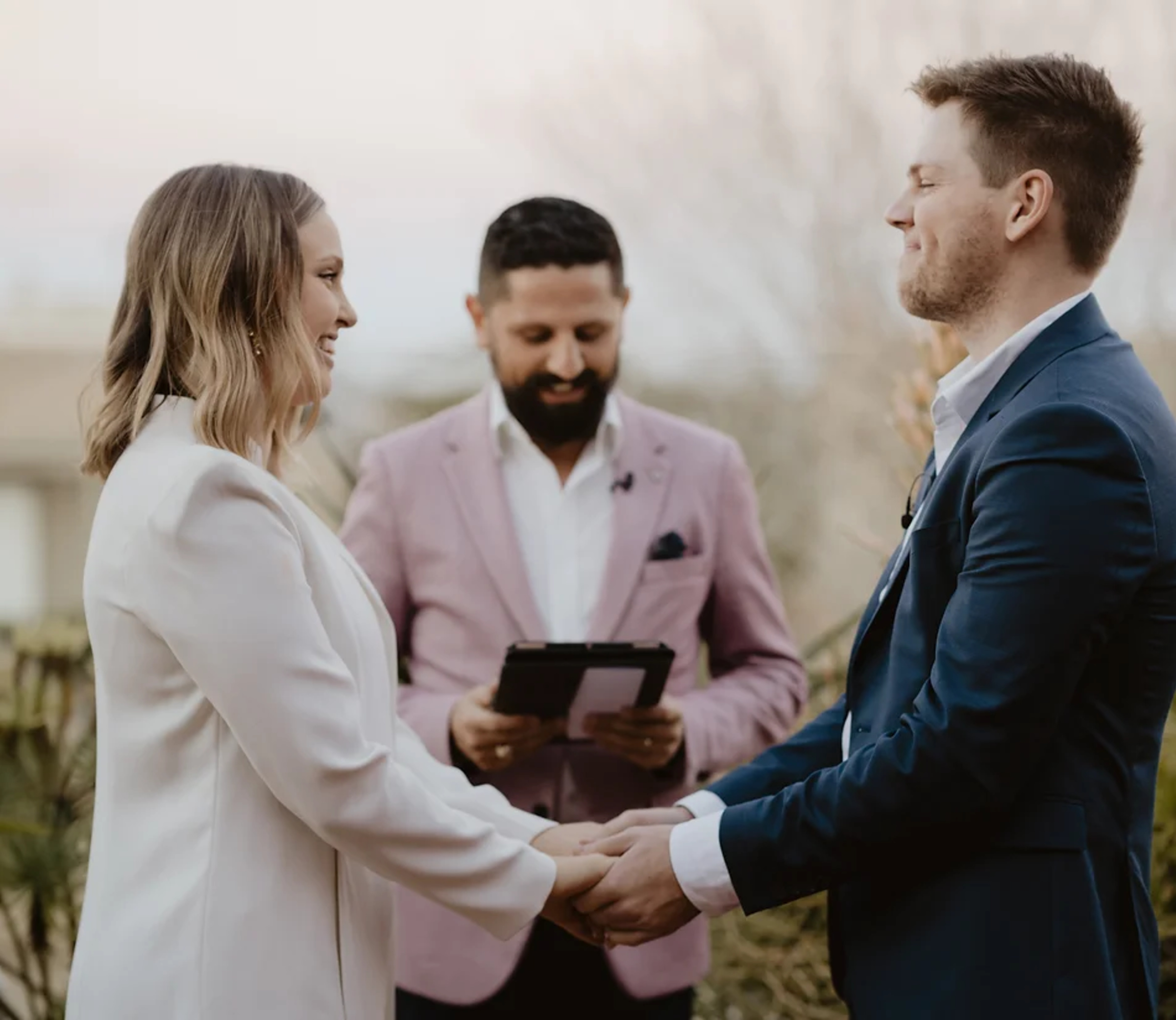 A couple holding hands and smiling during an outdoor wedding ceremony with an officiant standing behind them.