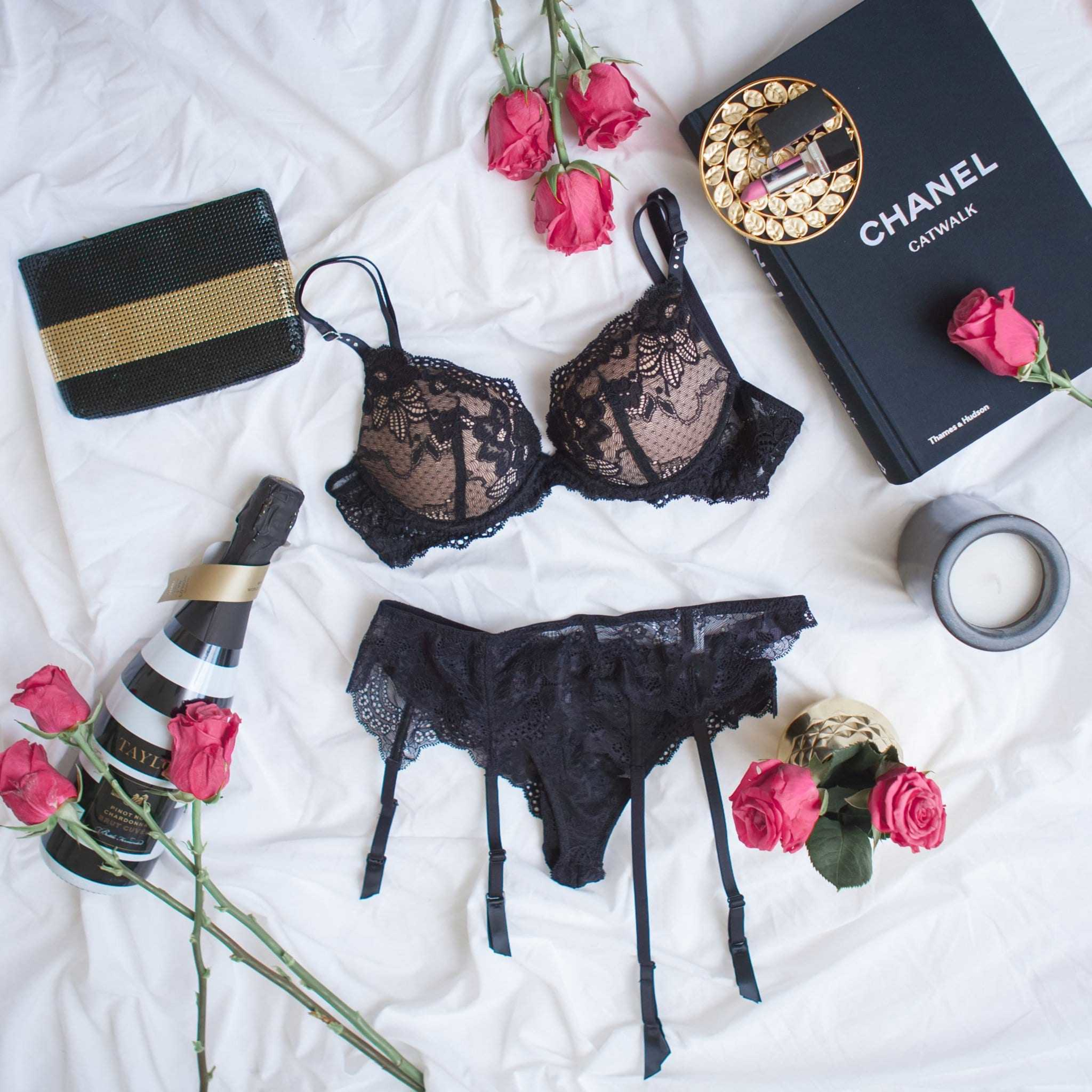 Flatlay of black lace lingerie with roses, champagne, and luxury accessories on a white bed.