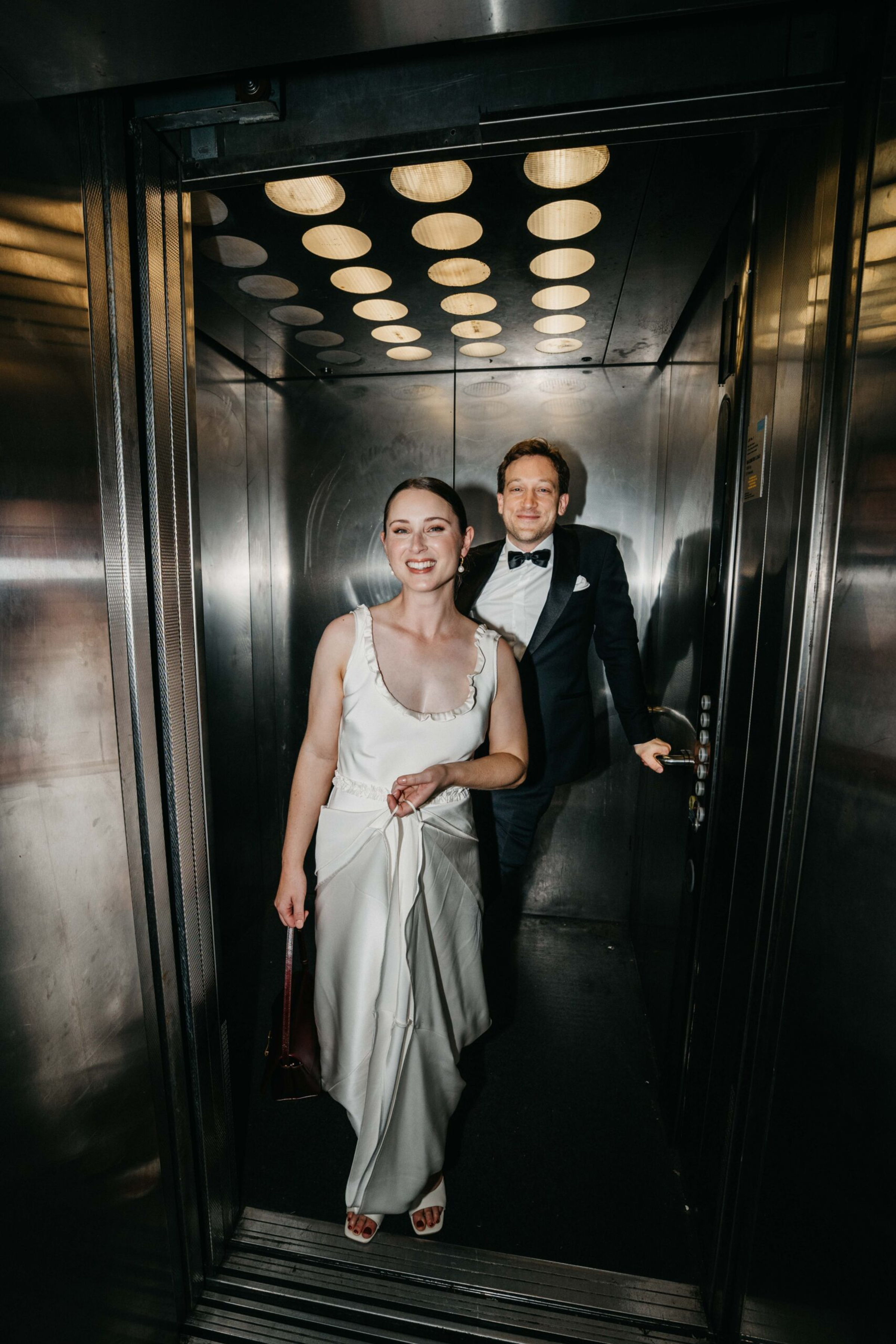 Bride and groom step out of a stainless steel elevator in modern wedding attire.