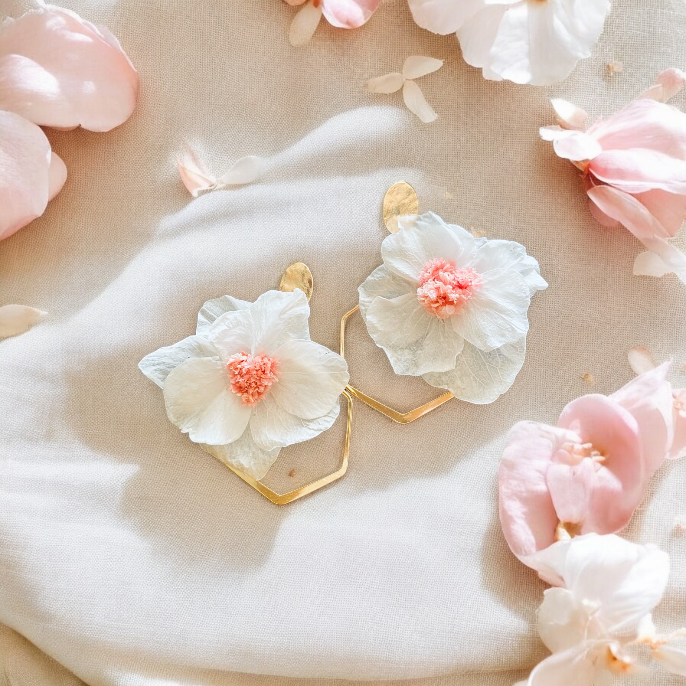 Delicate gold earrings with white and blush flower details surrounded by soft pink petals on fabric.