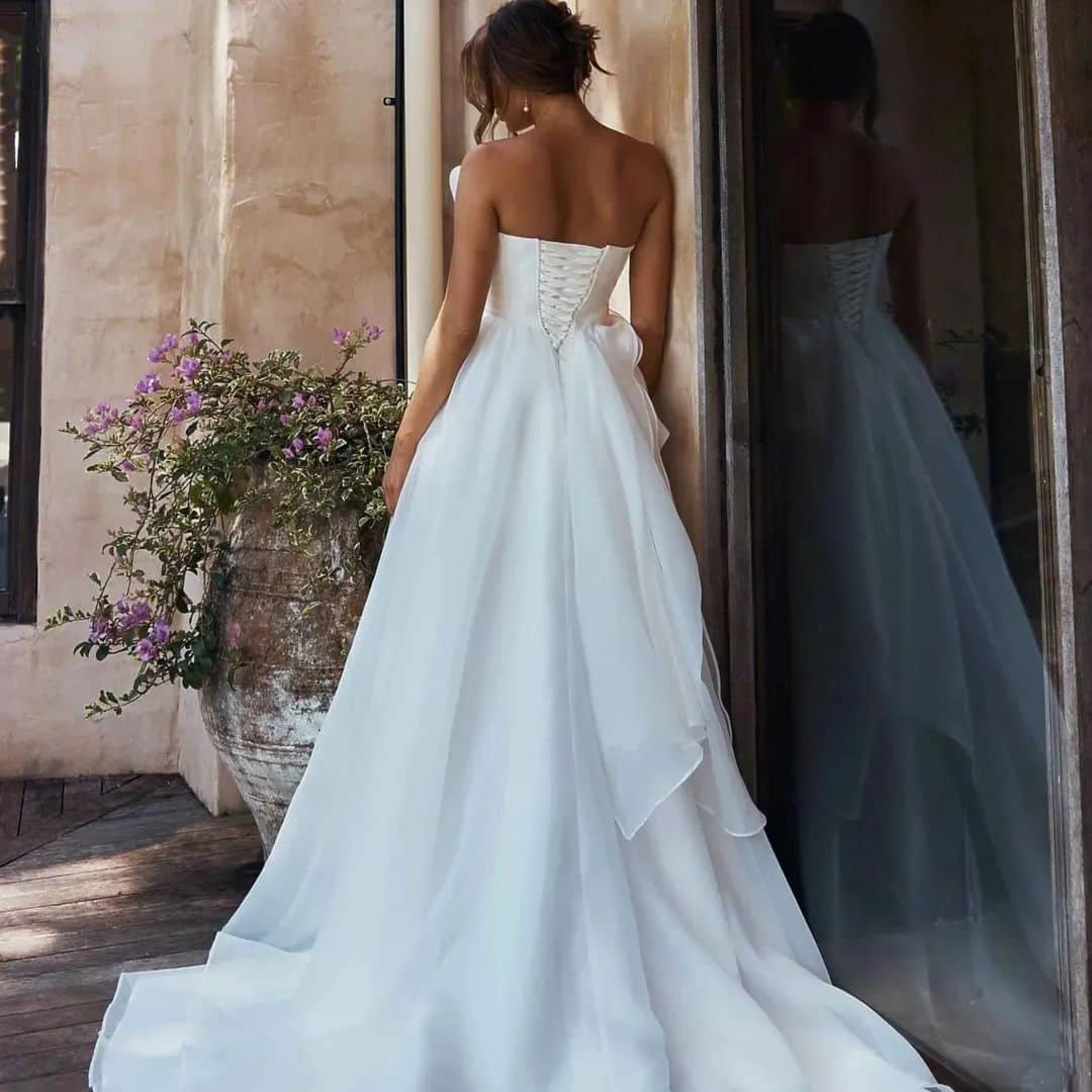 Bride in a strapless white gown stands by a rustic wall and large flower pot, showing the flowing train from behind.