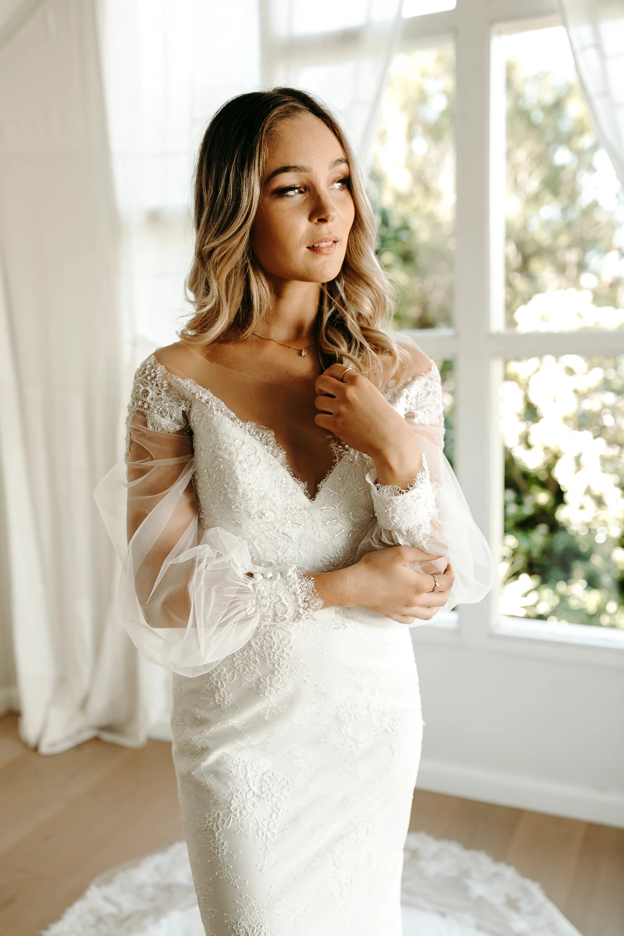 Bride in a lace long-sleeve wedding dress standing by a bright window with soft natural light.