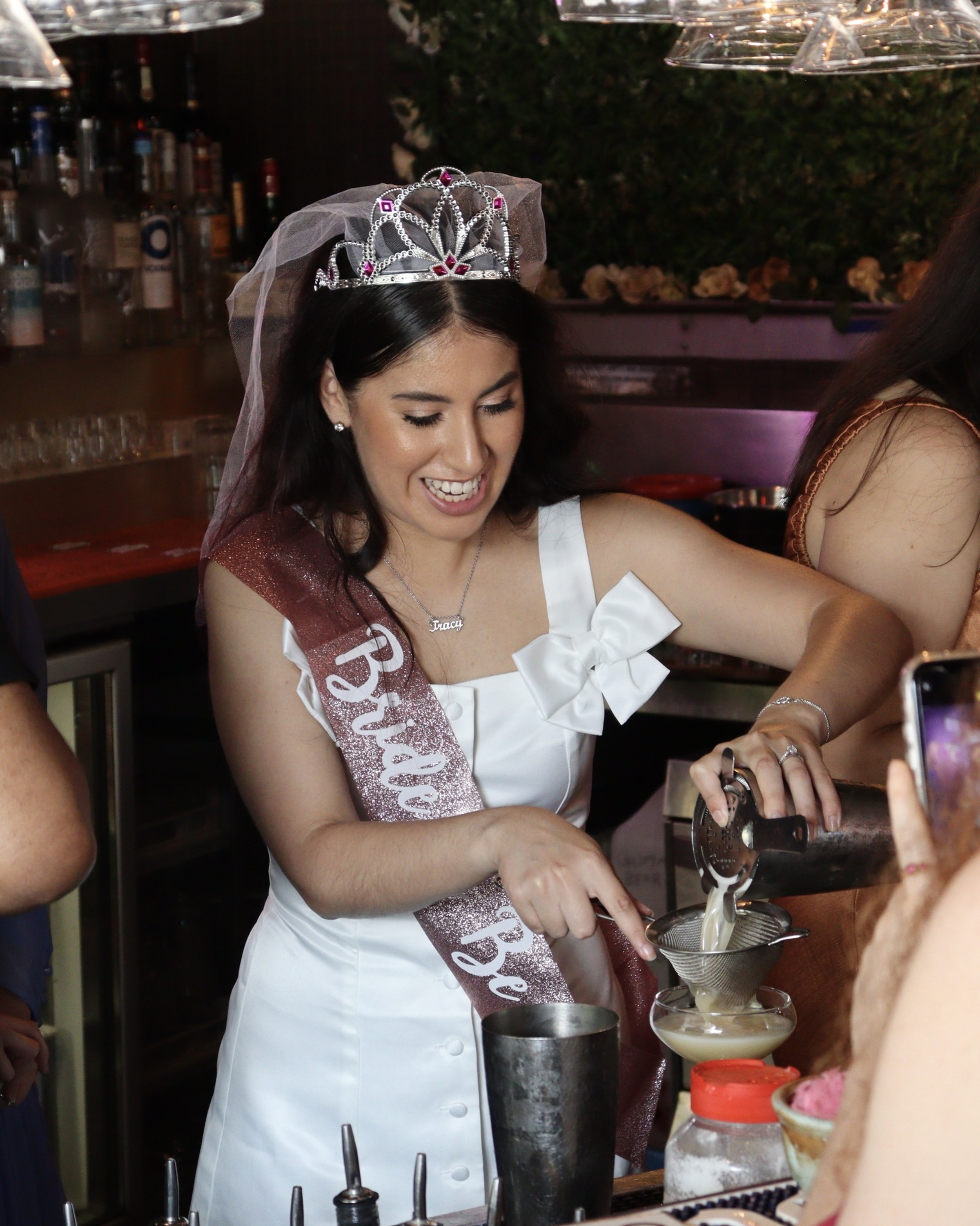 Bride-to-be wearing a tiara and sash pours cocktails at a lively bar during a pre-wedding celebration.