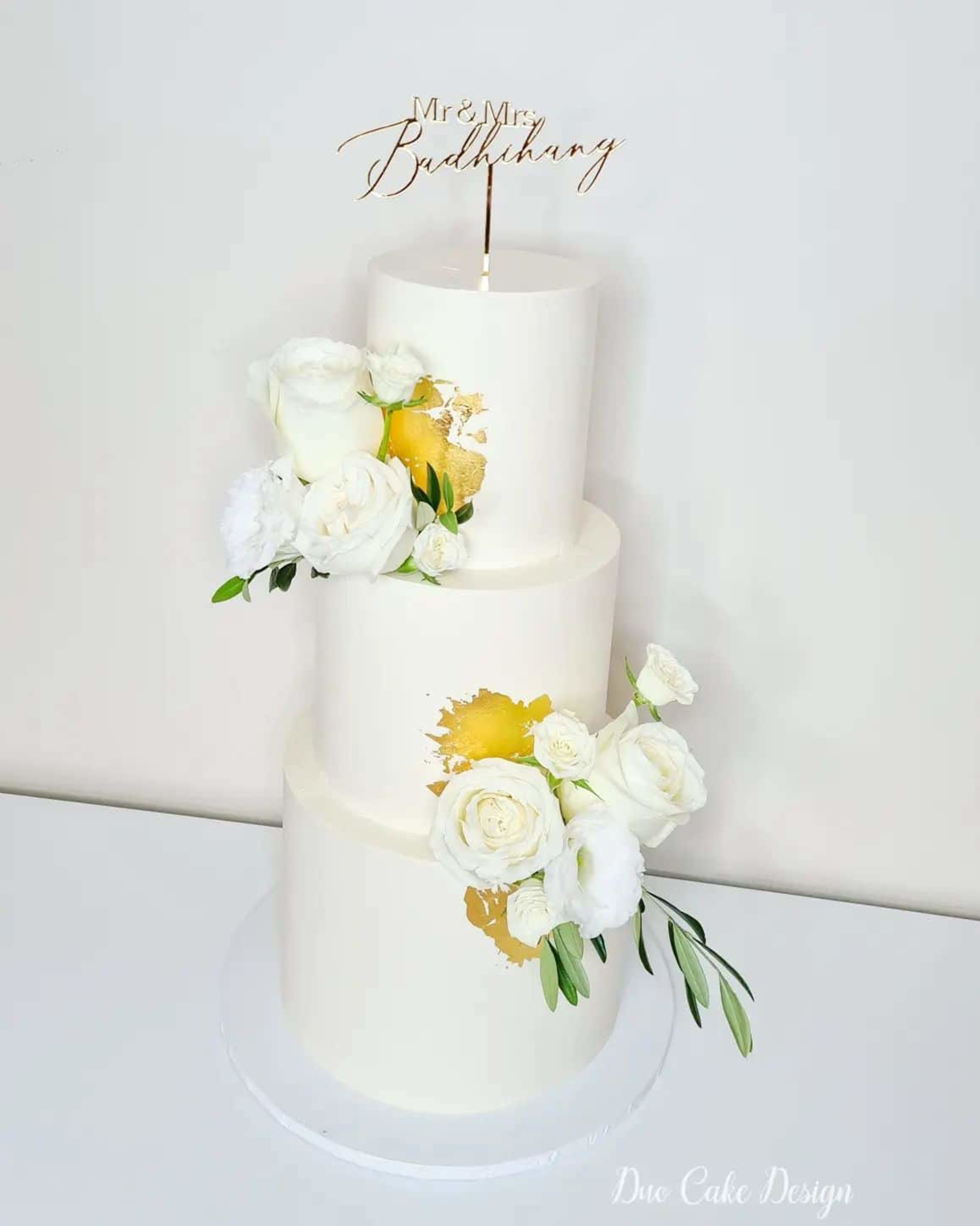 Three-tier white wedding cake with gold leaf accents, fresh white flowers, and a script Mr and Mrs topper.