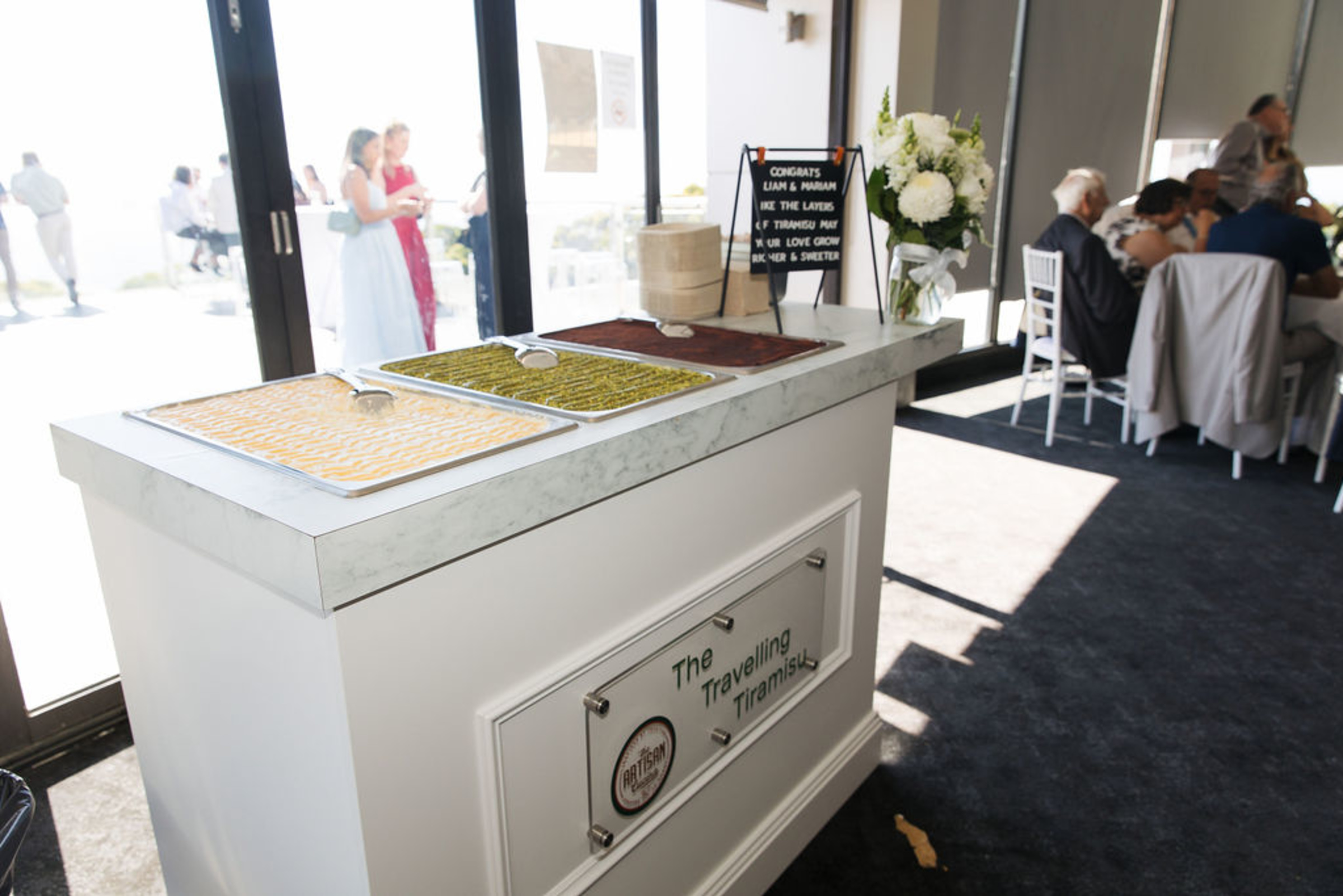 Wedding reception dessert station with trays of tiramisu on a white catering cart near seated guests.