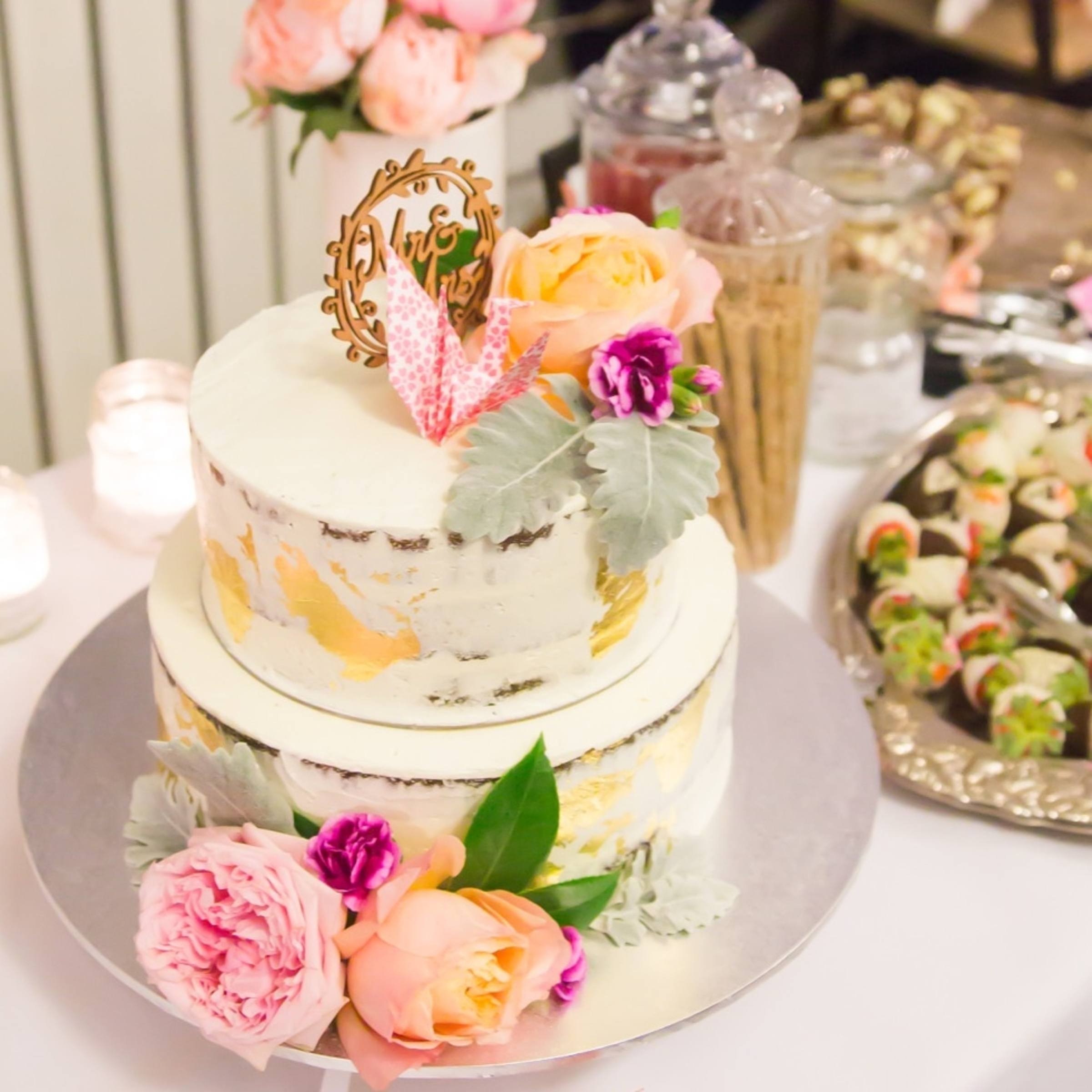 Two-tier white buttercream wedding cake with gold leaf and pink flowers on a dessert table.