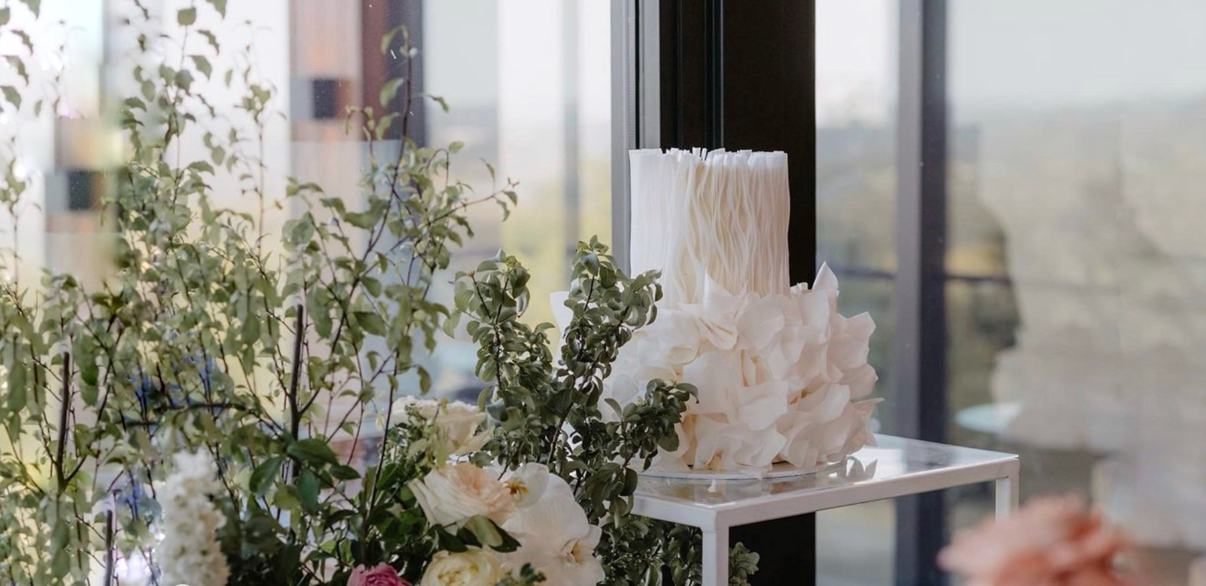 Tall white ruffled wedding cake displayed among lush greenery and pastel floral arrangements by a large window.