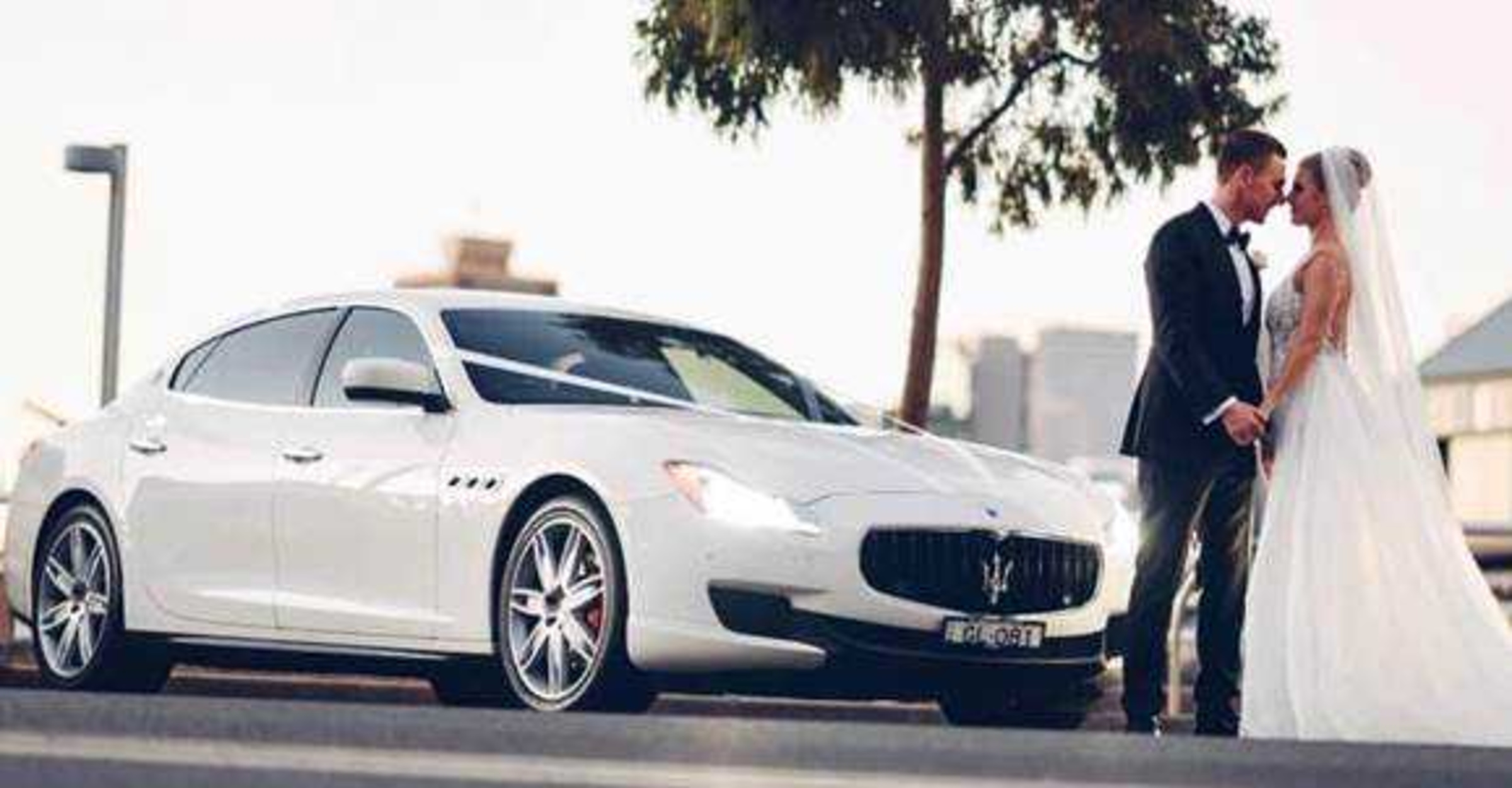 Bride and groom share a moment beside a white luxury wedding car on a city street.