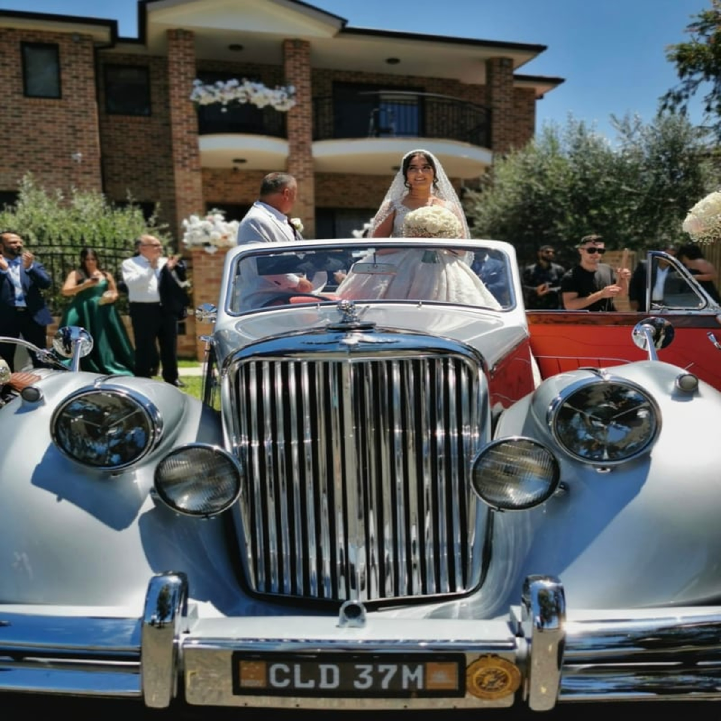 Bride in a classic silver convertible wedding car outside a brick house surrounded by guests.