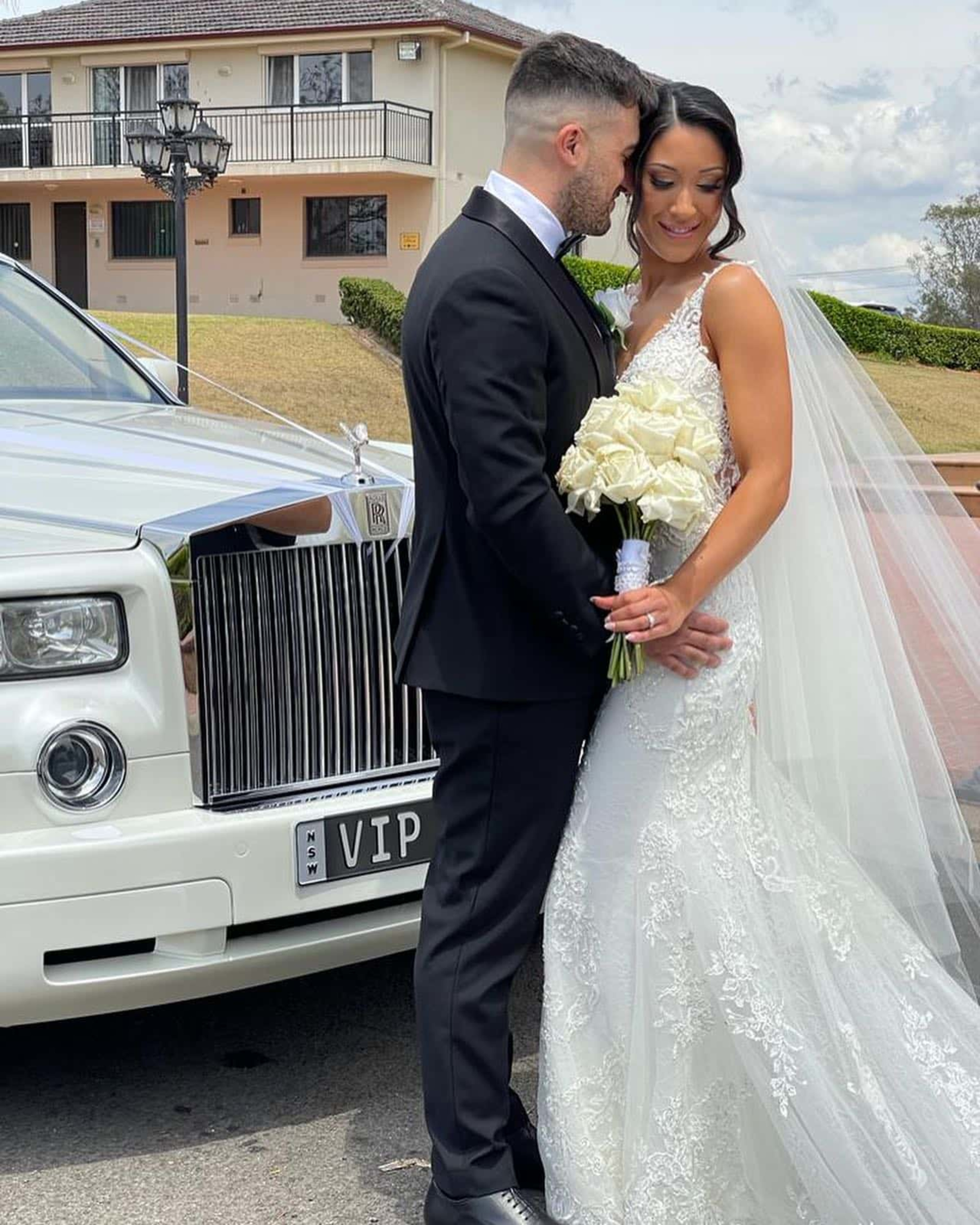 Bride and groom embrace beside a white luxury wedding car, with the bride holding a bouquet of white roses.