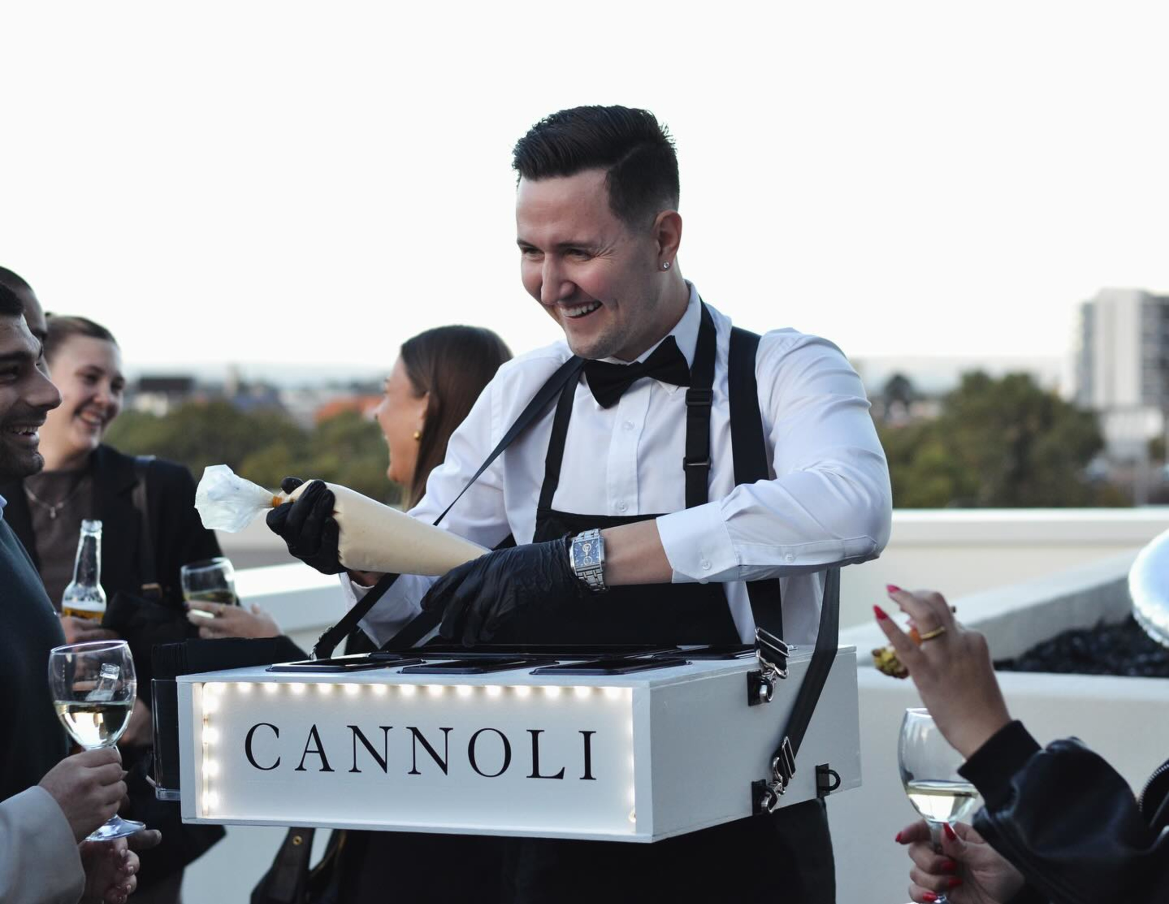 Smiling server fills cannoli from a portable dessert box for guests at an outdoor wedding reception.