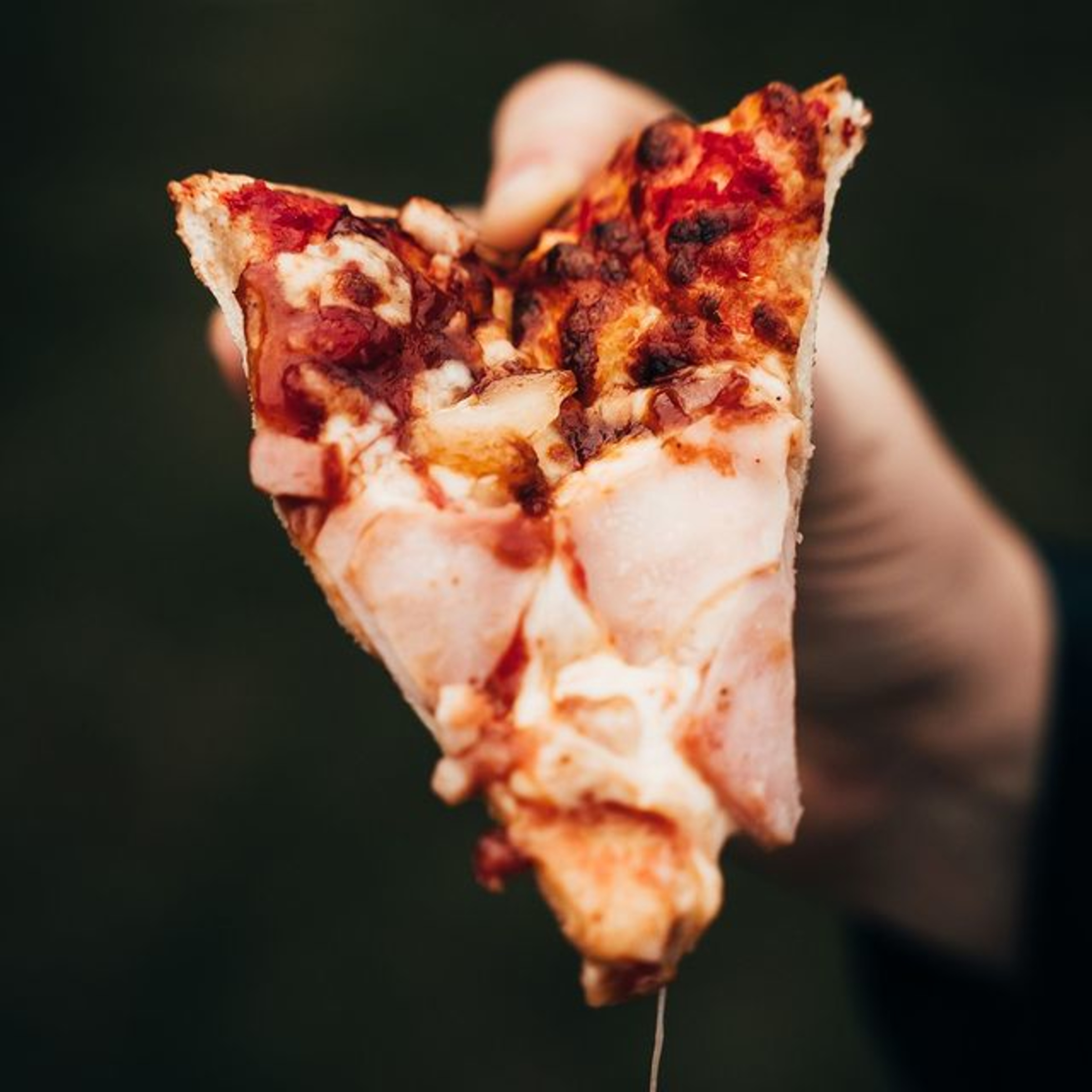 Close-up of a hand holding a cheesy slice of pizza against a dark blurred background.