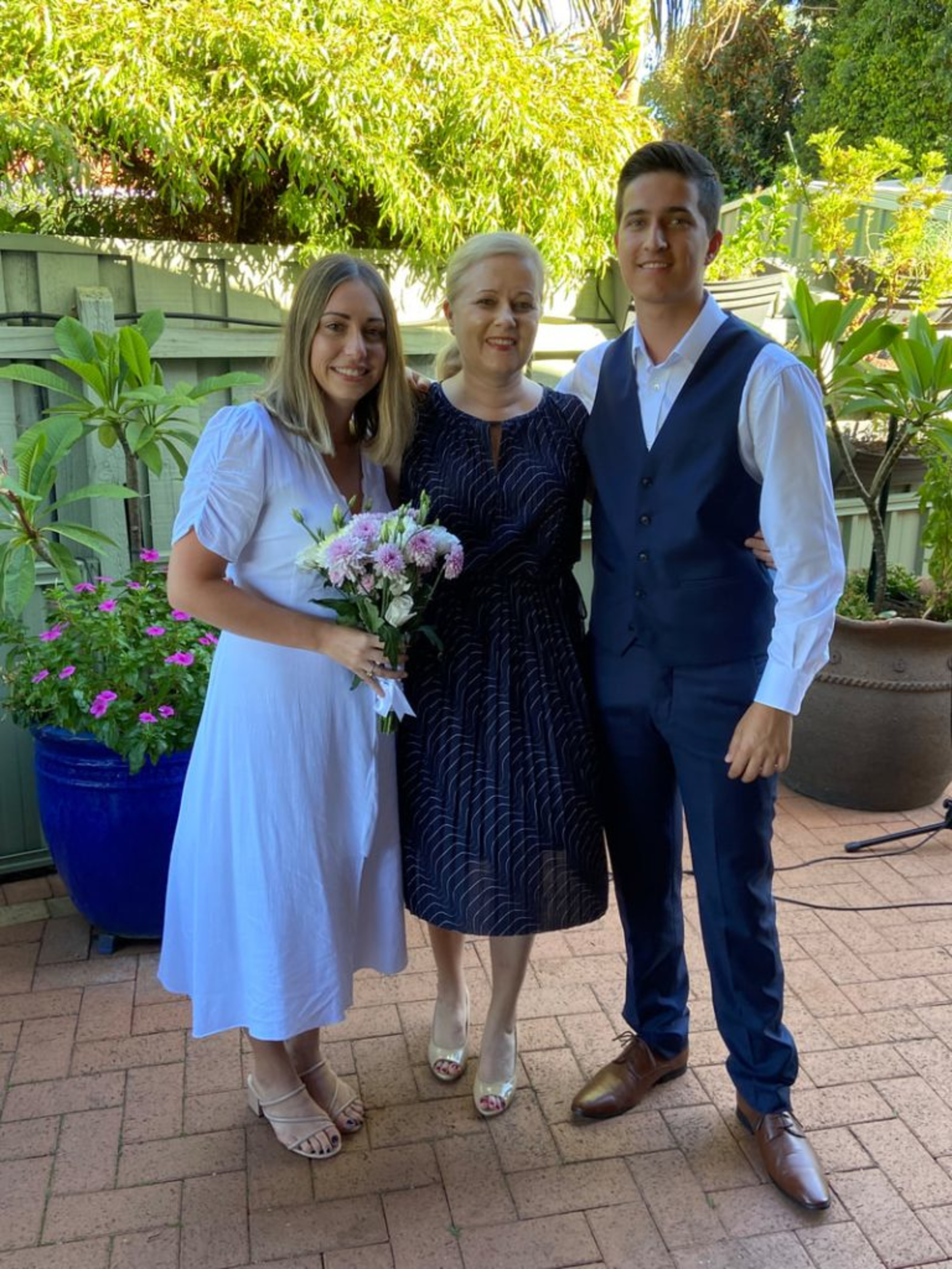 Bride and groom stand with their celebrant in a sunny garden setting holding a pink and white bouquet.