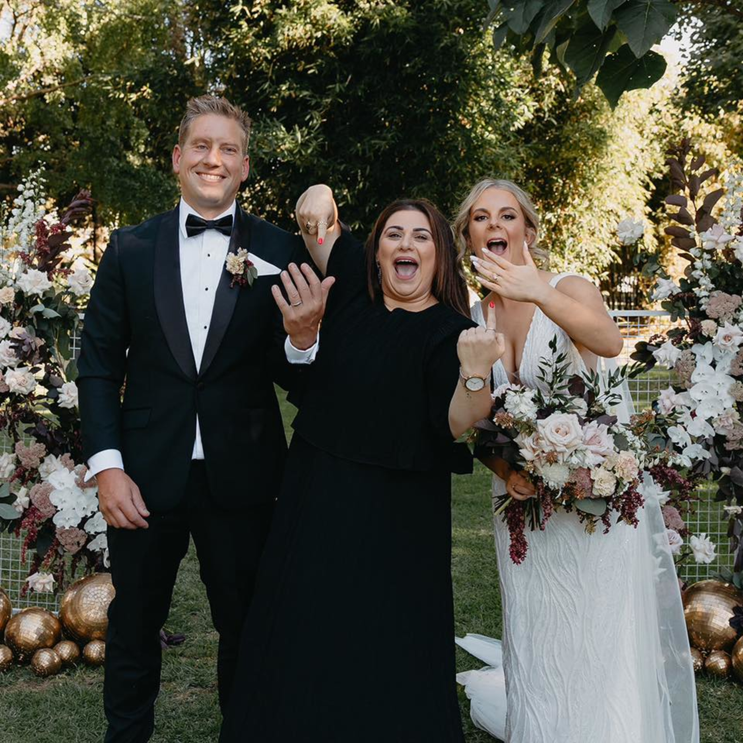 Joyful bride, groom, and celebrant posing outdoors in front of lush floral arrangements showing off wedding rings.