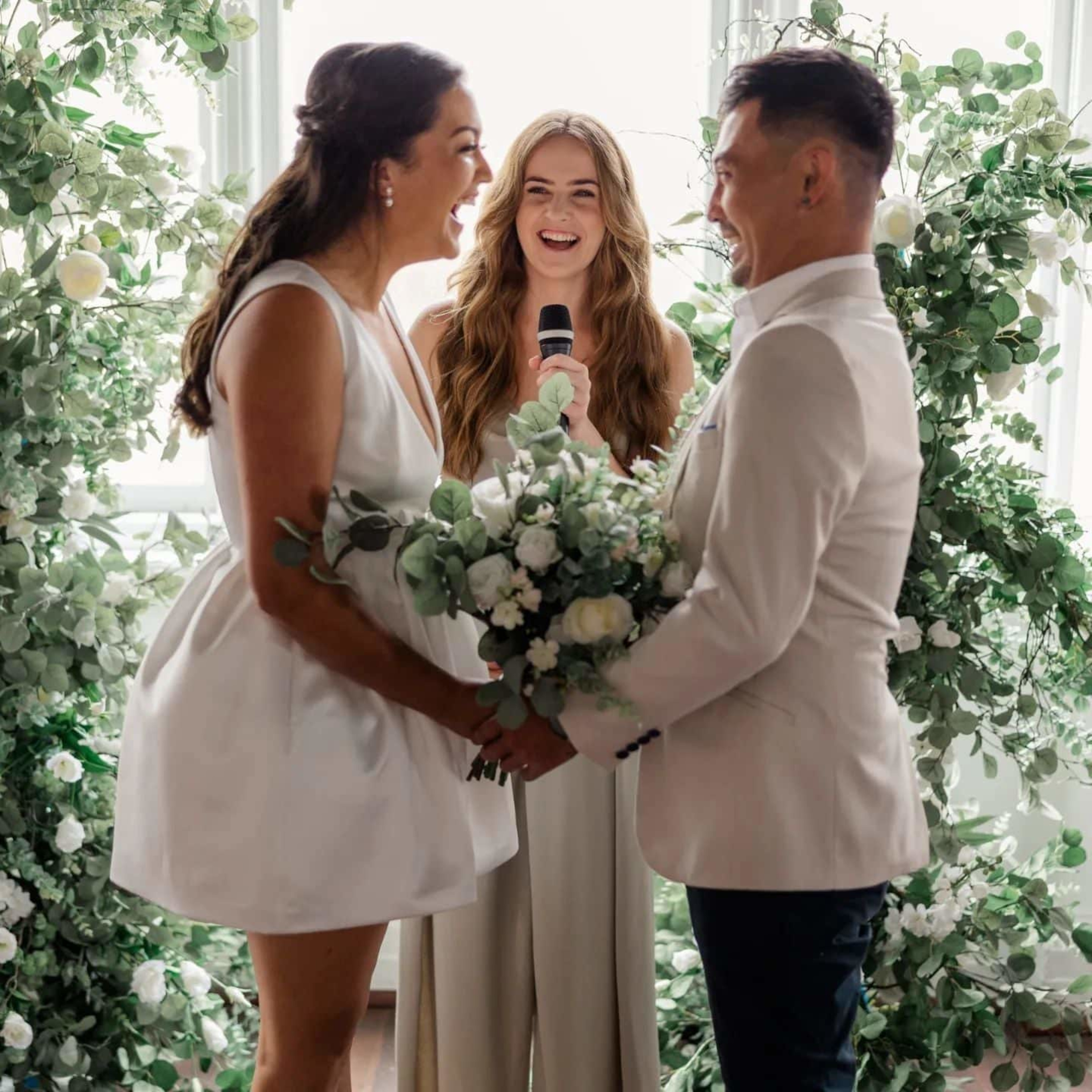 Joyful couple holding hands during an indoor wedding ceremony with lush greenery and a smiling officiant.