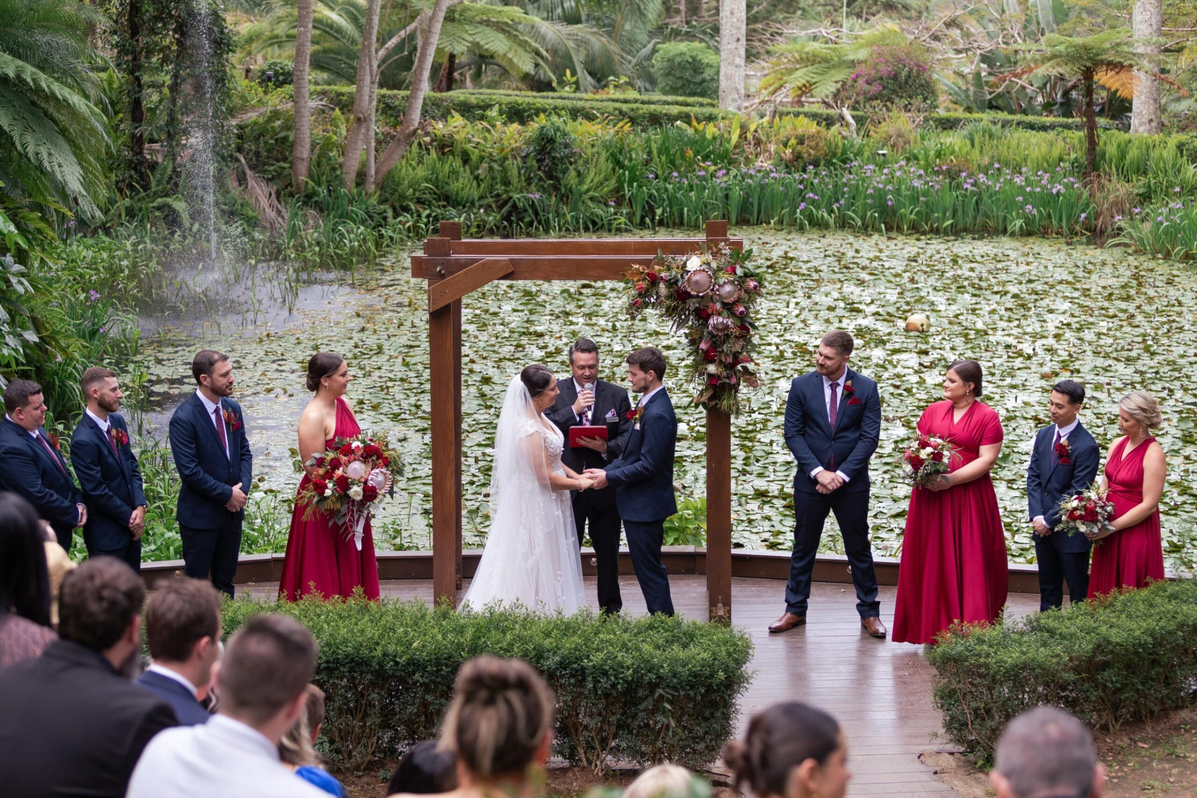 Outdoor wedding ceremony by a lily pond with the couple under a wooden floral arbor and bridal party in red dresses and navy suits.