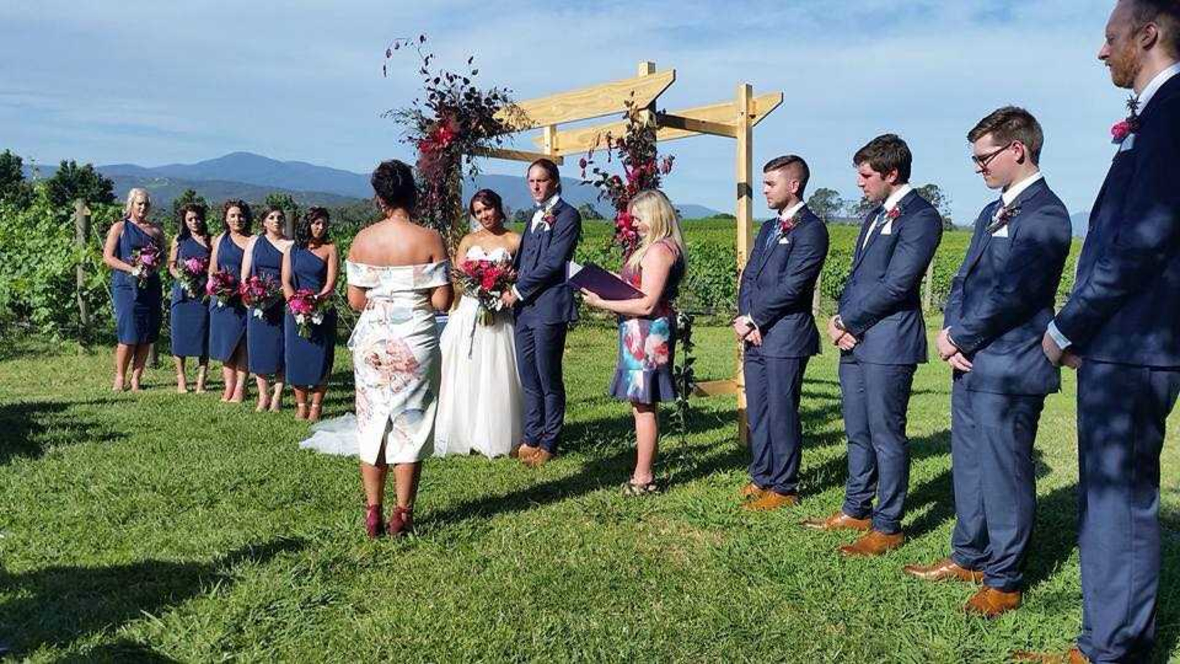 Outdoor vineyard wedding ceremony with the couple and bridal party standing beneath a floral wooden arch.