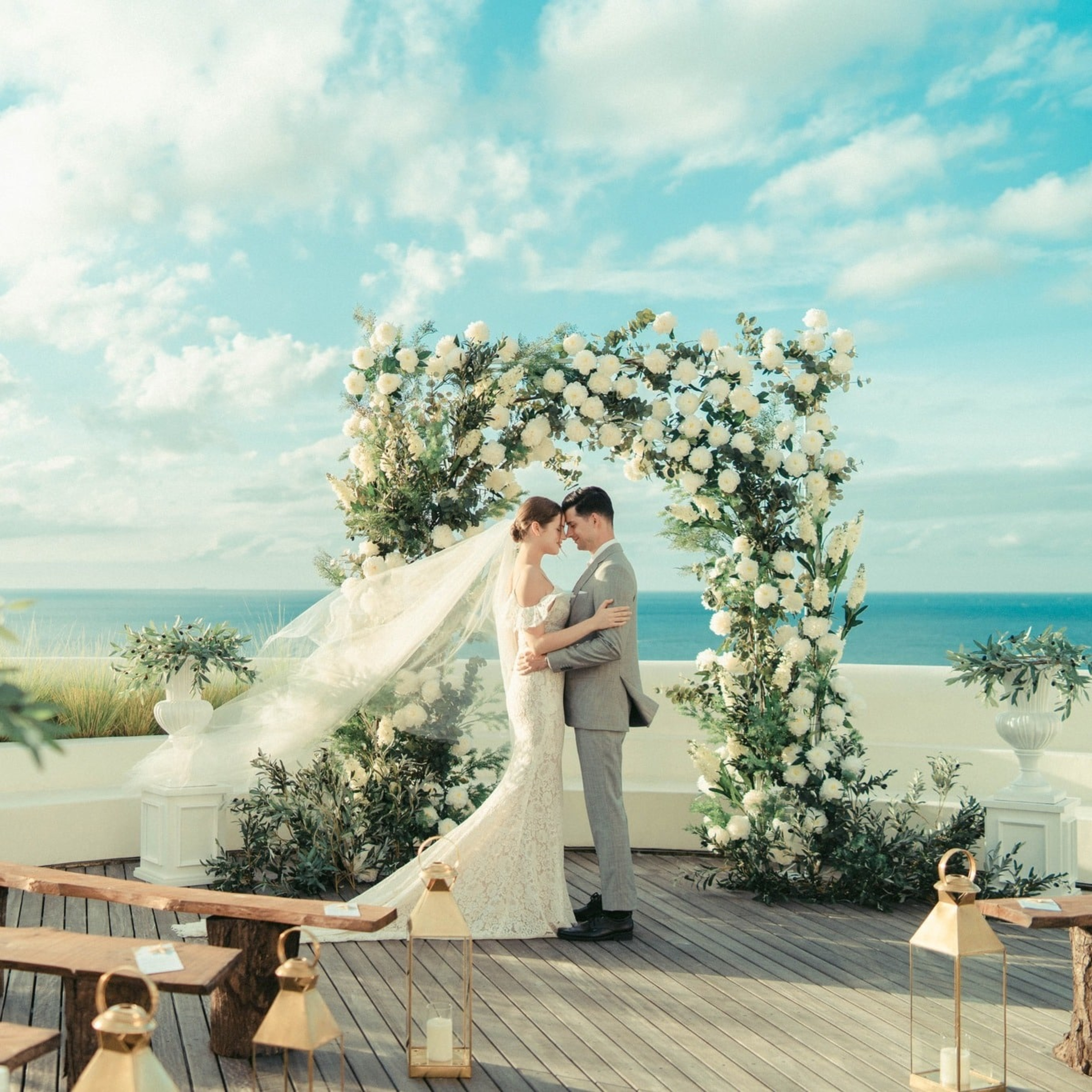 Bride and groom embrace under a lush floral arch at an elegant seaside wedding ceremony.