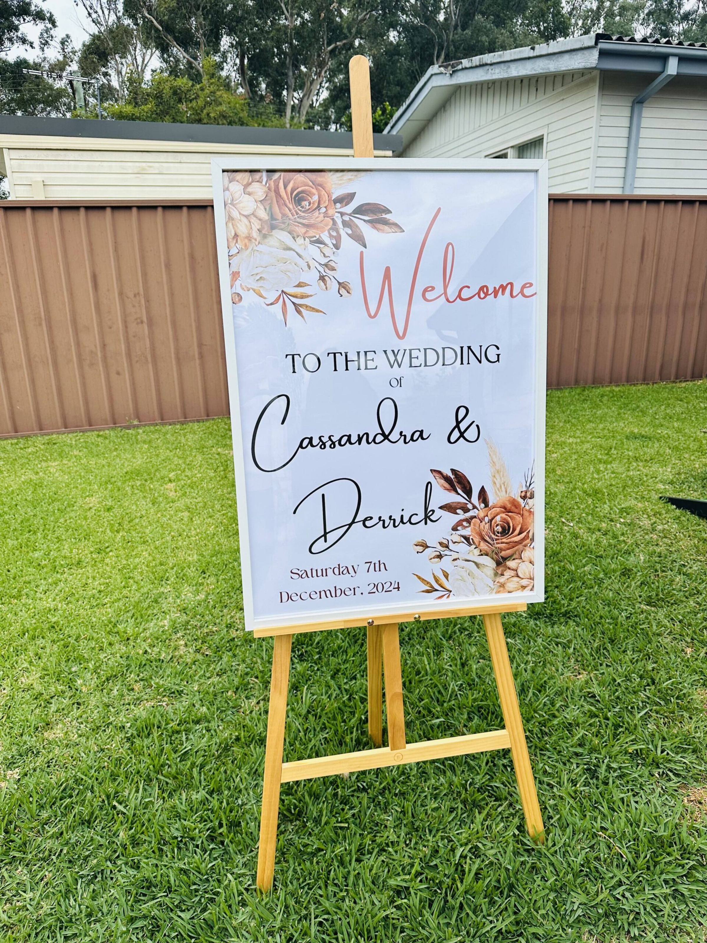 Outdoor wedding welcome sign on wooden easel with floral design and calligraphy text on the lawn.